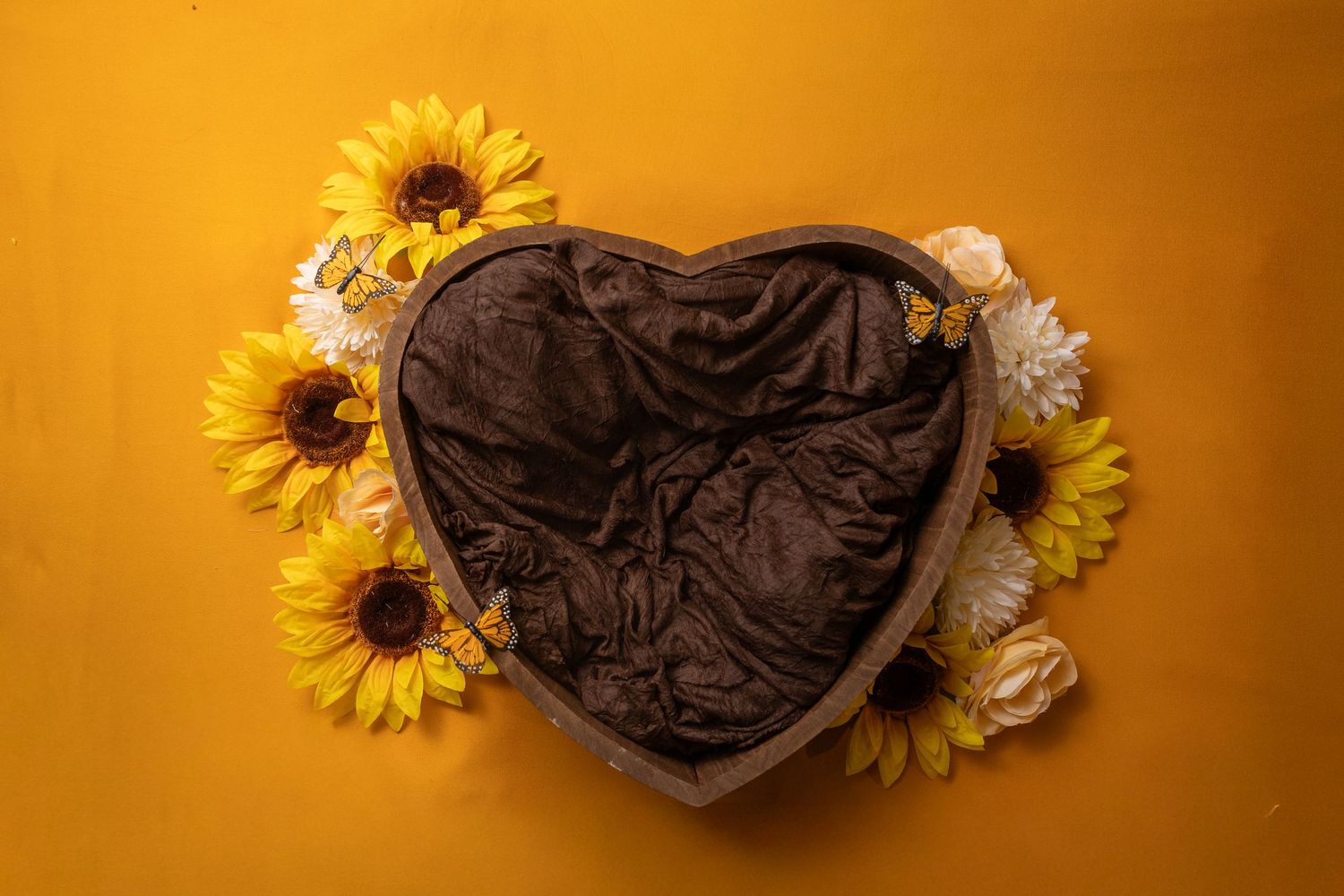 Heart-shaped brown velvet box surrounded by yellow sunflowers and white chrysanthemums on a golden yellow background.