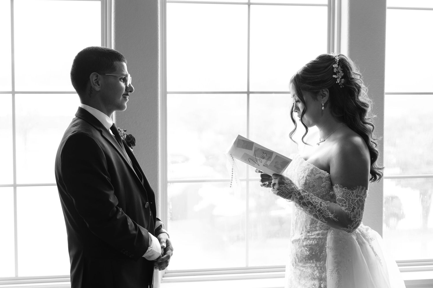 Black and white photo of bride reading vows to groom by bright windows in elegant wedding attire.
