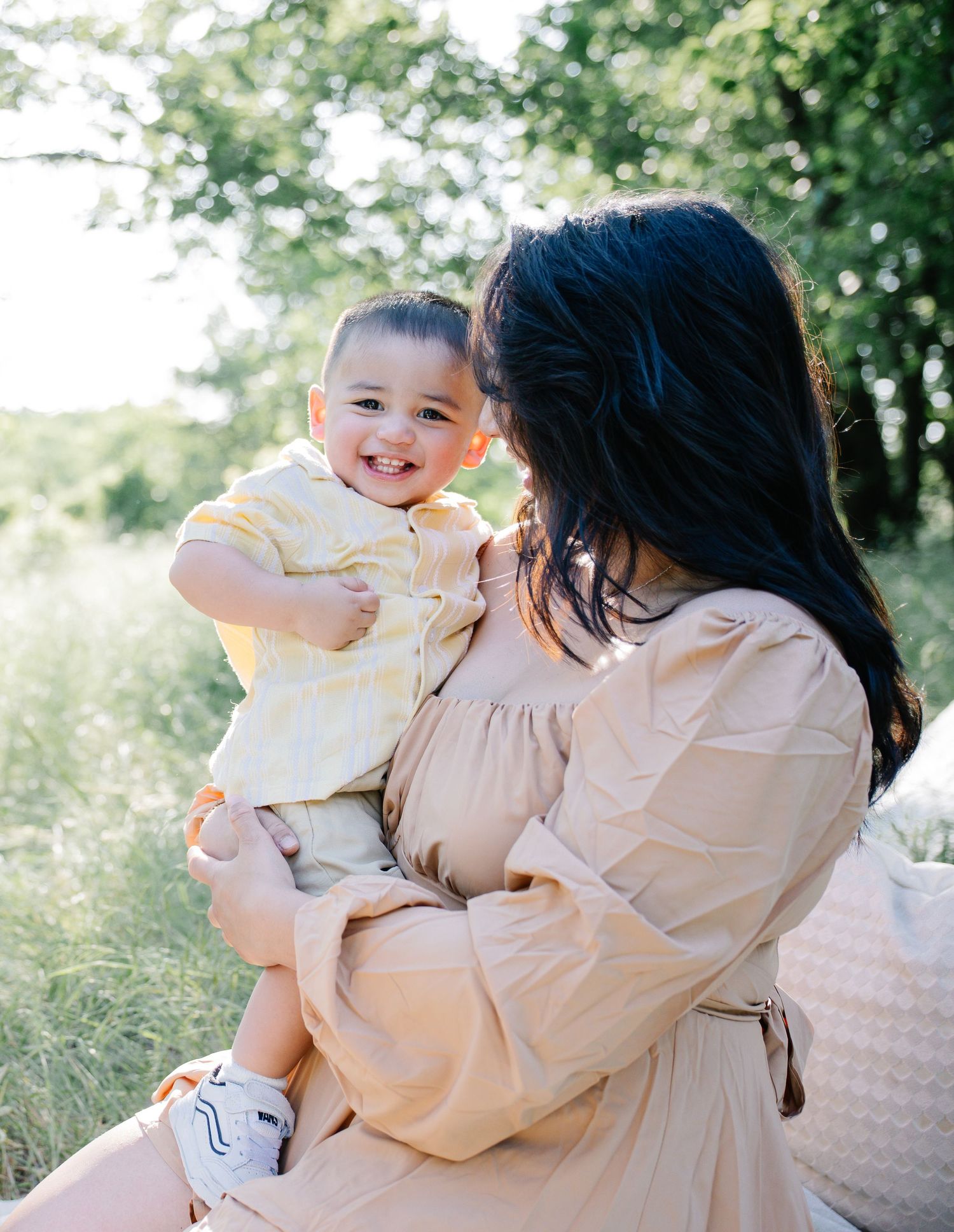 A smiling baby in a yellow outfit being held by a dark-haired woman in a beige dress outdoors.