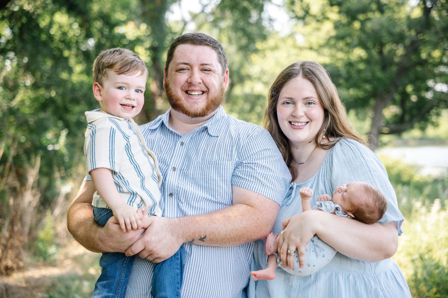 Happy family of four posing outdoors, parents holding toddler and newborn baby, wearing coordinating blue outfits.