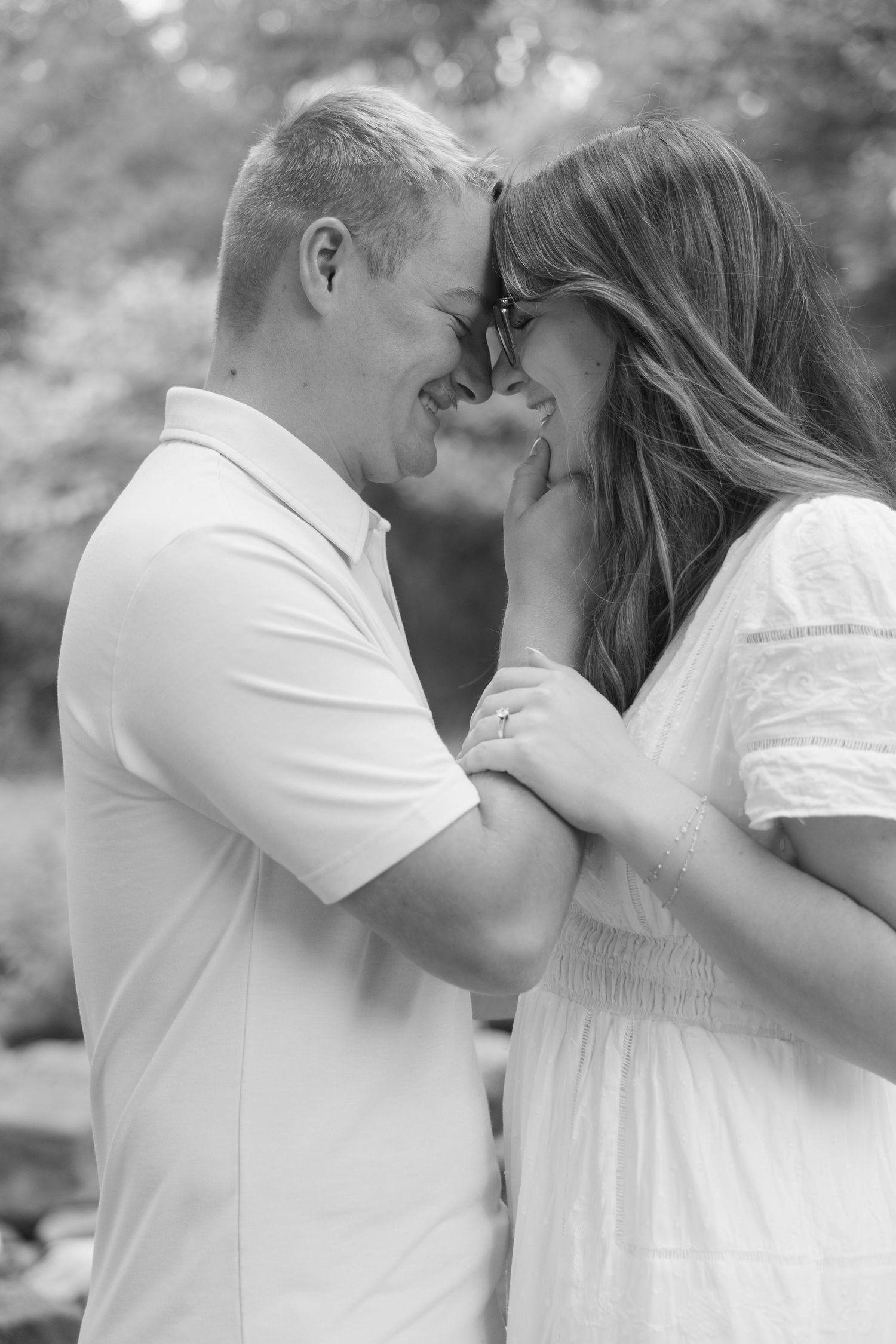 Black and white photo of a romantic couple touching foreheads and holding hands outdoors.