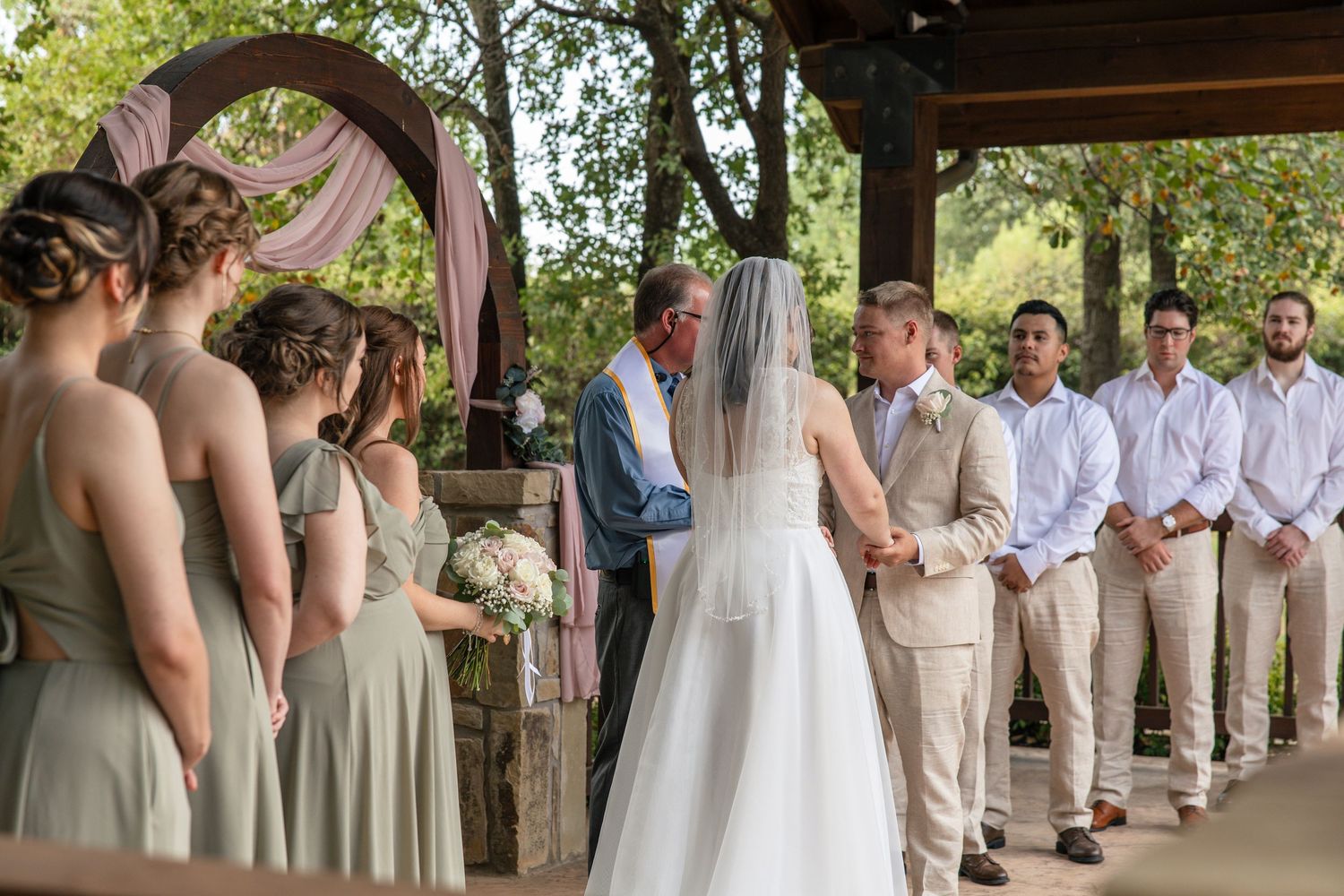 Bride in a white gown and groom exchange vows under a draped wooden arch, with bridesmaids and groomsmen looking on.