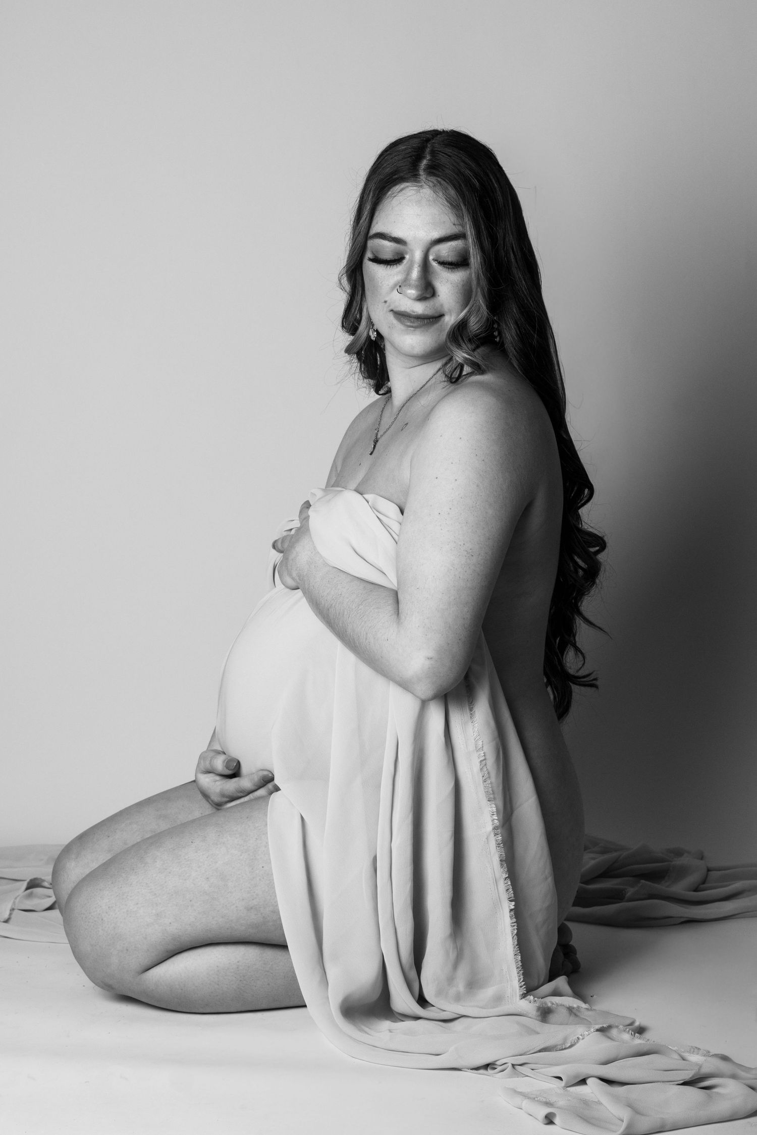 Black and white maternity photo of a pregnant woman kneeling, cradling her belly, wearing a draped fabric dress.