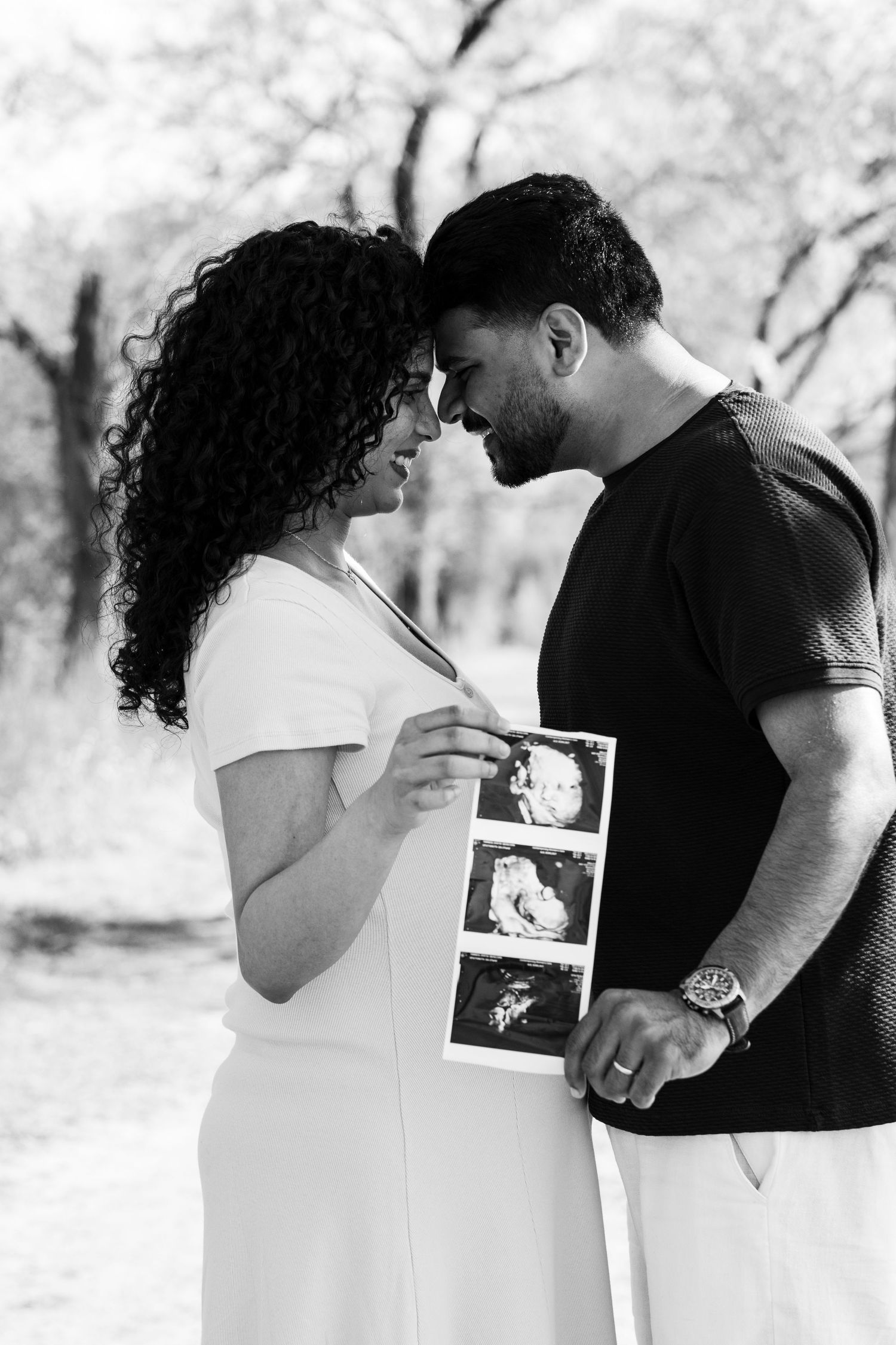 Black and white photo of expecting couple touching foreheads while holding ultrasound images in park.