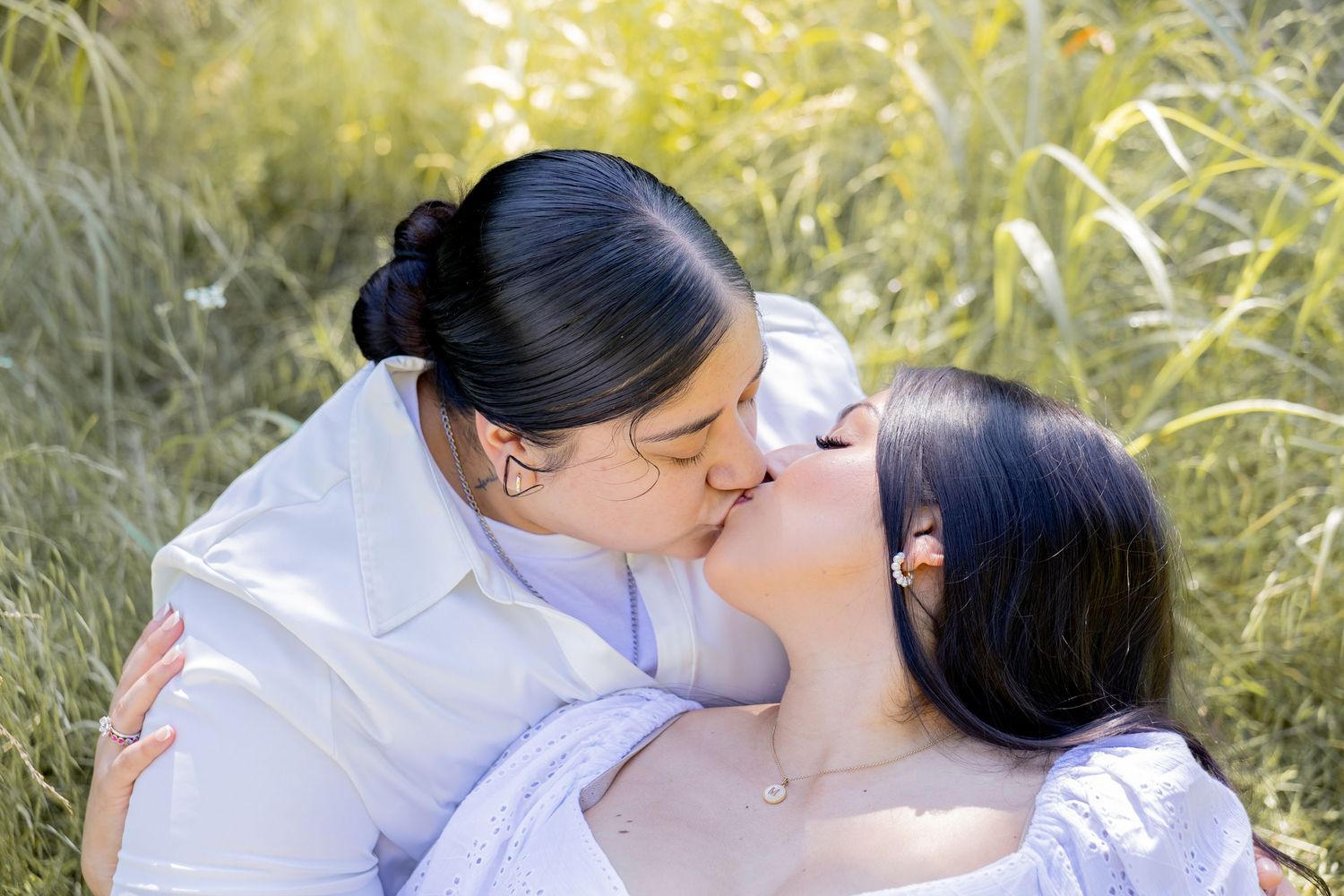 Two women share a romantic kiss while sitting together in a sunlit field of tall green grass.