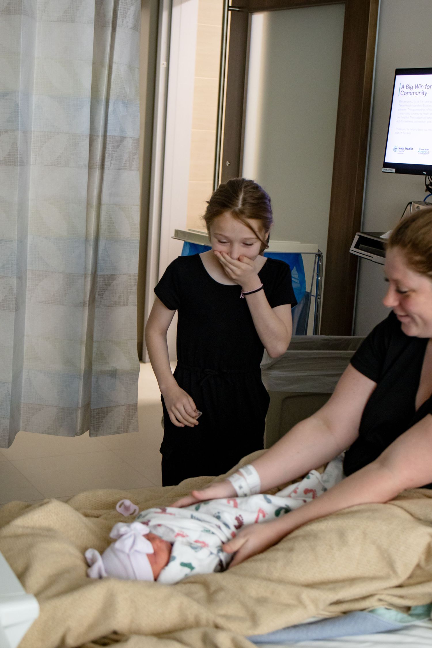 Emotional young girl covering her mouth meeting her newborn sibling in a hospital room.