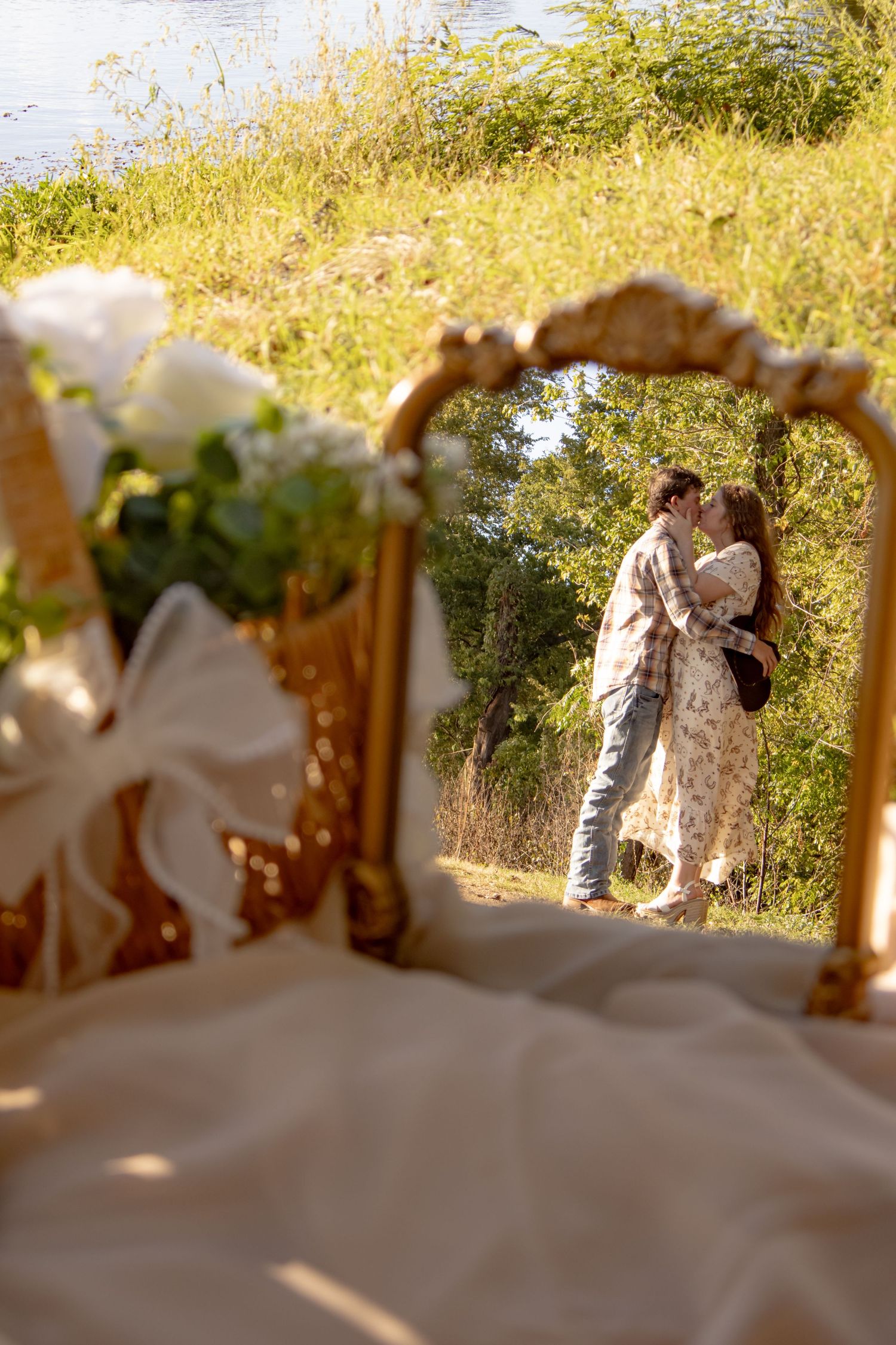 Couple kissing near a river reflected in a gold ornate mirror with a white rose bouquet and bow in the foreground.