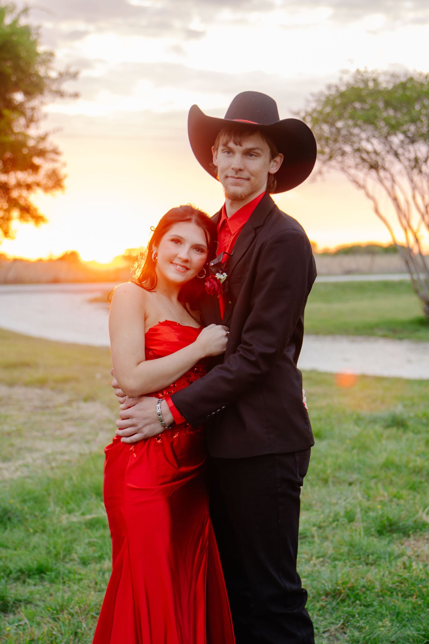 Prom couple posing at sunset, girl in red strapless gown, boy in black suit and cowboy hat.