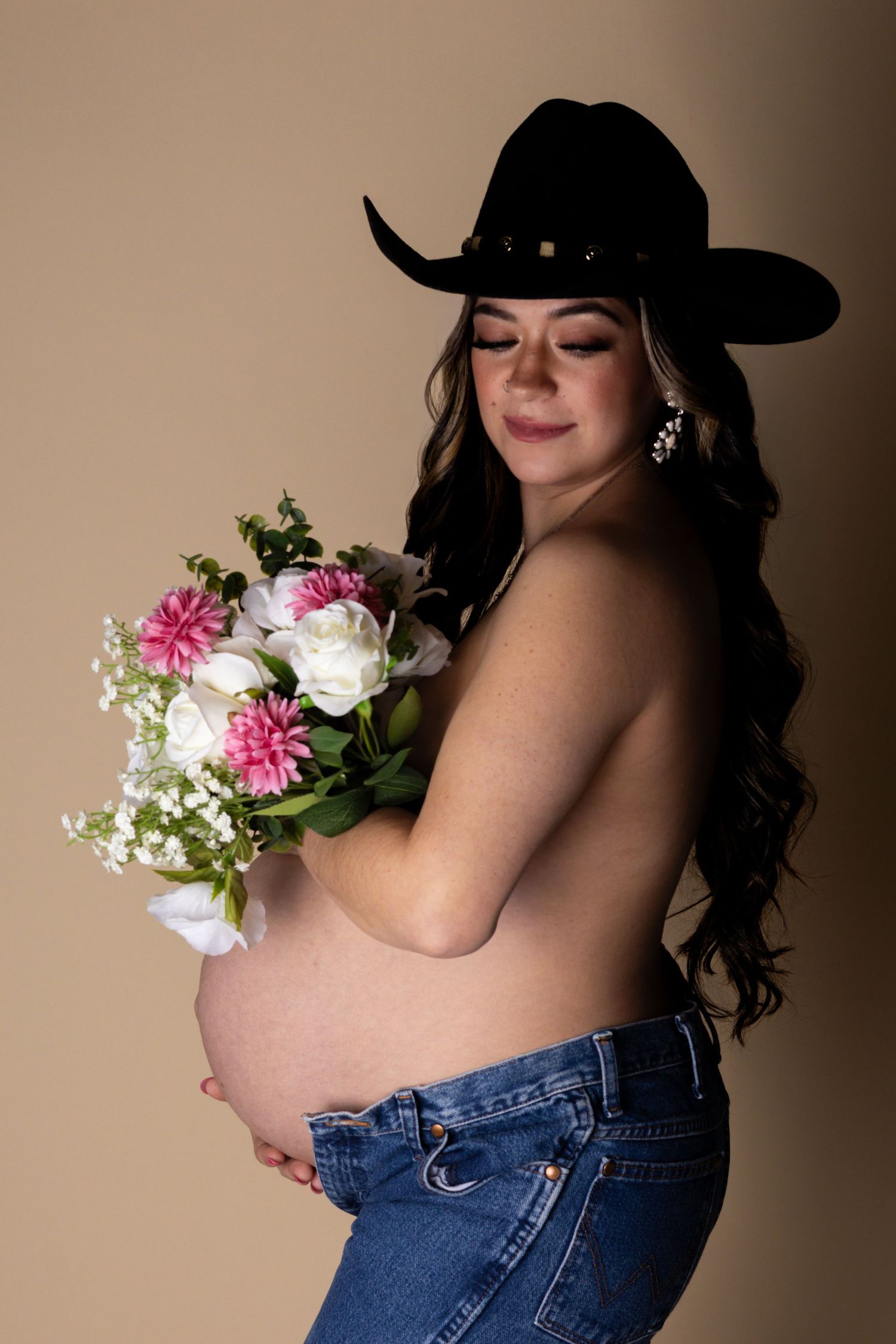 Pregnant woman in black cowboy hat holding pink and white flower bouquet in studio.