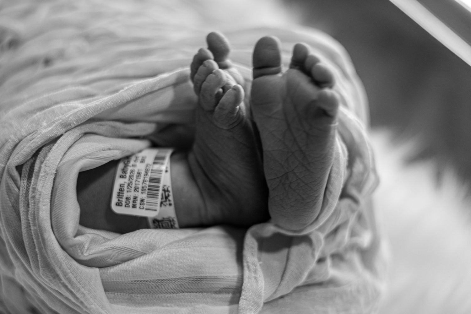 Newborn baby feet with hospital ID bracelet wrapped in blanket, black and white.