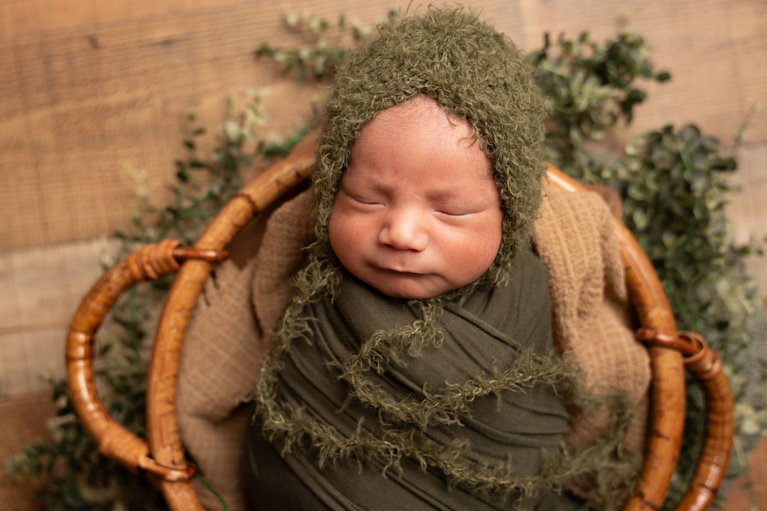 Sleeping newborn baby in an olive green knit bonnet and wrap, nestled in a rattan basket with eucalyptus greenery on a rustic wood backdrop.
