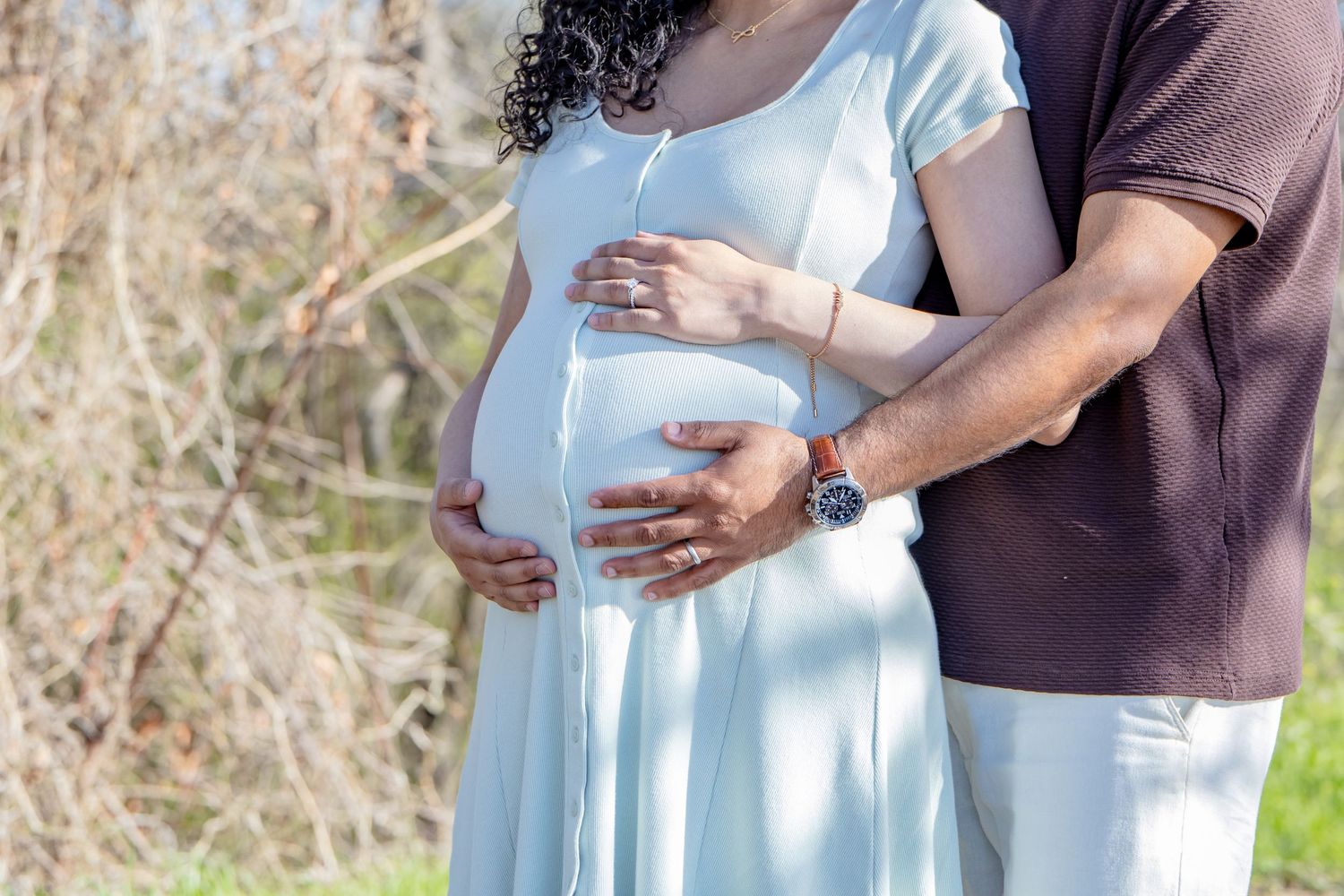 Expecting couple embracing in nature, both hands lovingly holding pregnant woman's baby bump outside.
