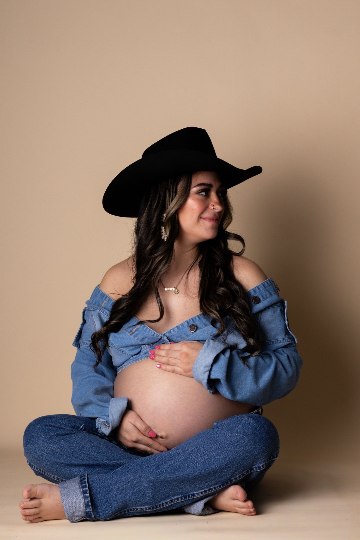 Pregnant woman in denim top and jeans with black cowboy hat smiling in a studio setting.