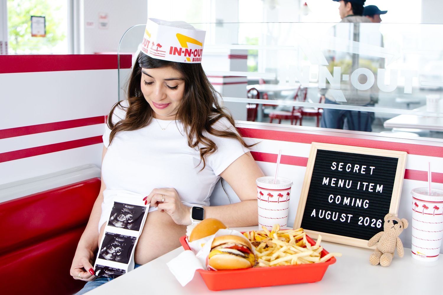 Pregnant woman at In-N-Out Burger holding ultrasound photos beside a tray of food and a baby announcement sign.