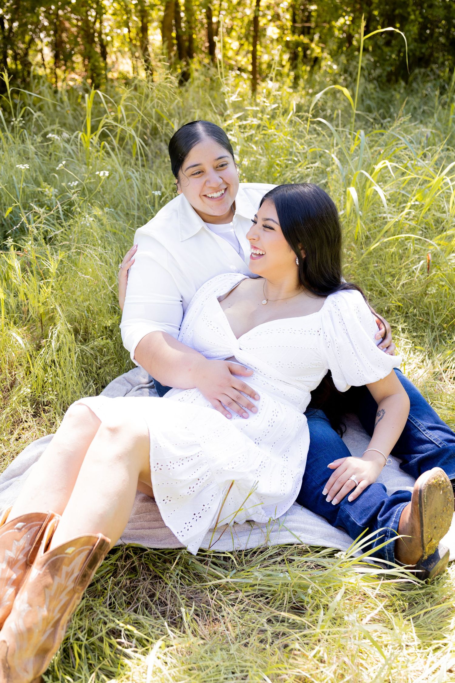 Two women laugh and cuddle together while sitting in tall grass with a scenic wooded background.