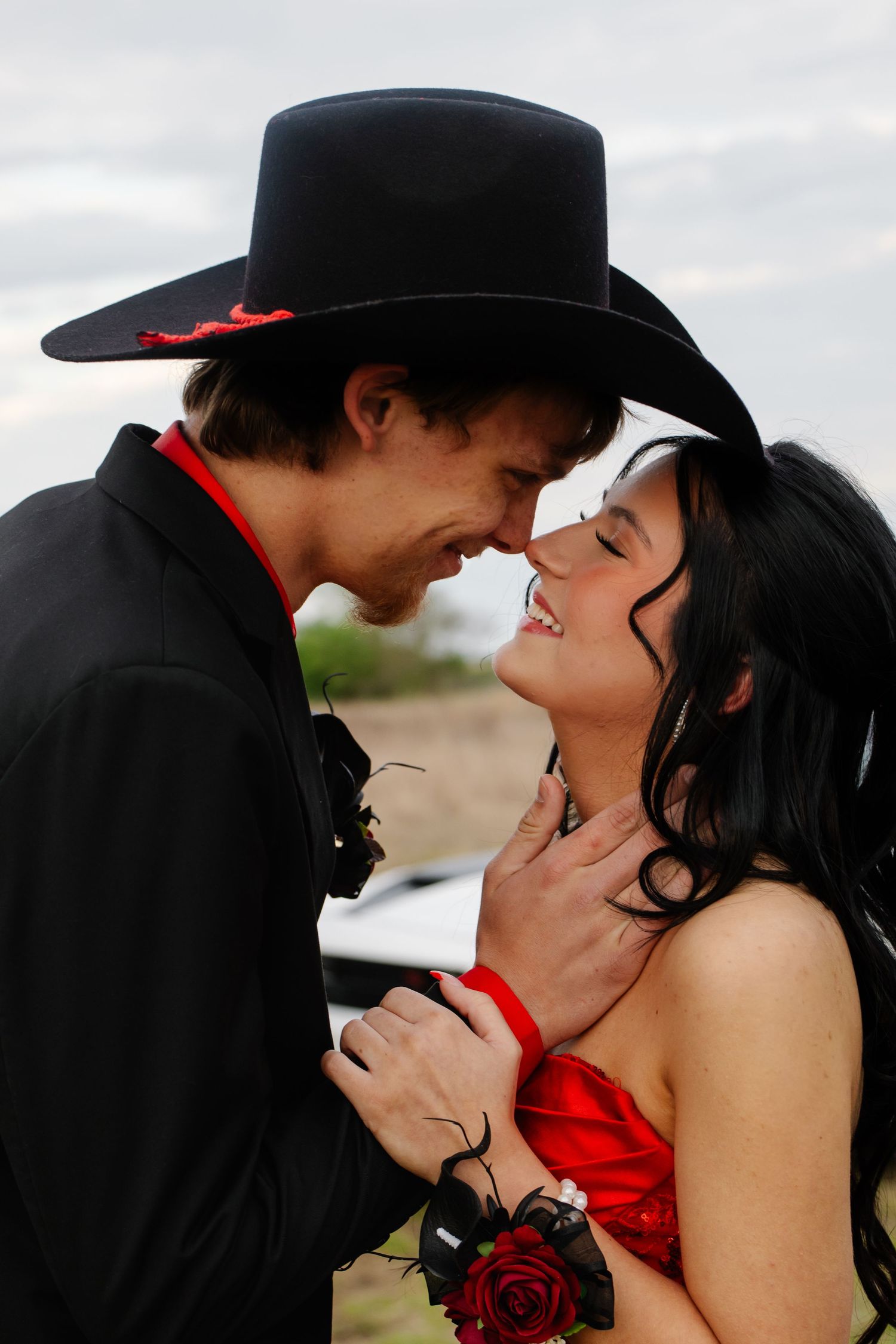 Young couple in prom attire, woman in red dress and man in black cowboy hat, sharing romantic moment.