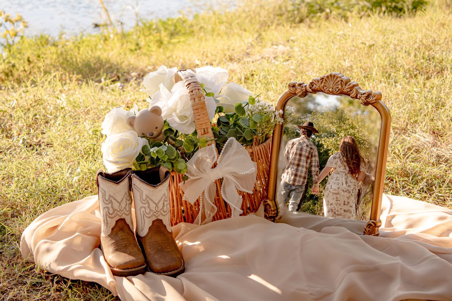 Rustic maternity flat lay with cowboy boots, white roses in a basket, and a mirror reflecting a couple outdoors.