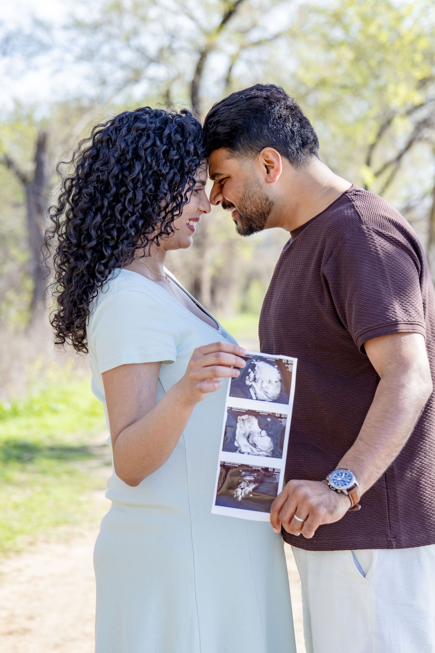 Happy pregnant couple sharing an intimate moment holding baby ultrasound images together in a sunlit park.