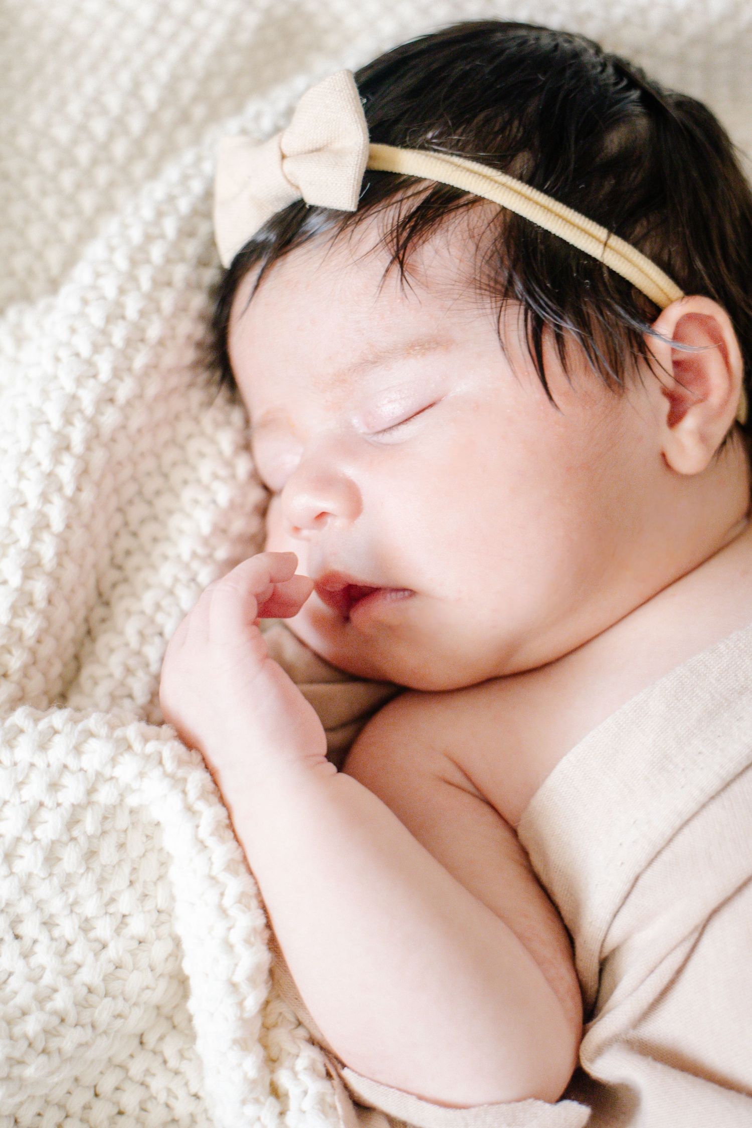 Close-up of sleeping newborn baby with dark hair wearing a small cream bow headband.
