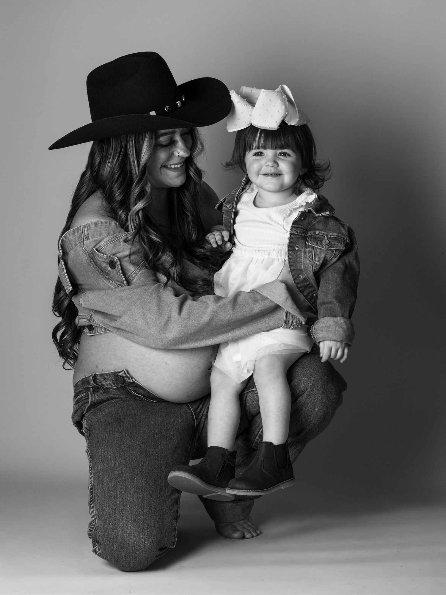 Black and white photo of a woman in a cowboy hat kneeling with a toddler girl in a white dress and bow.