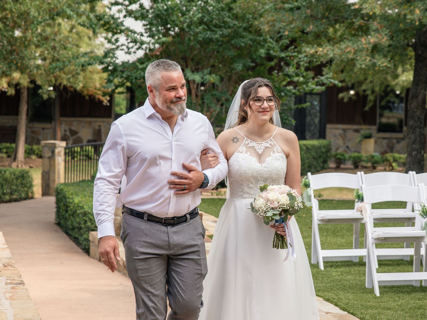 Father in a white dress shirt walks a smiling bride in a lace wedding gown holding a bouquet down an outdoor garden aisle.