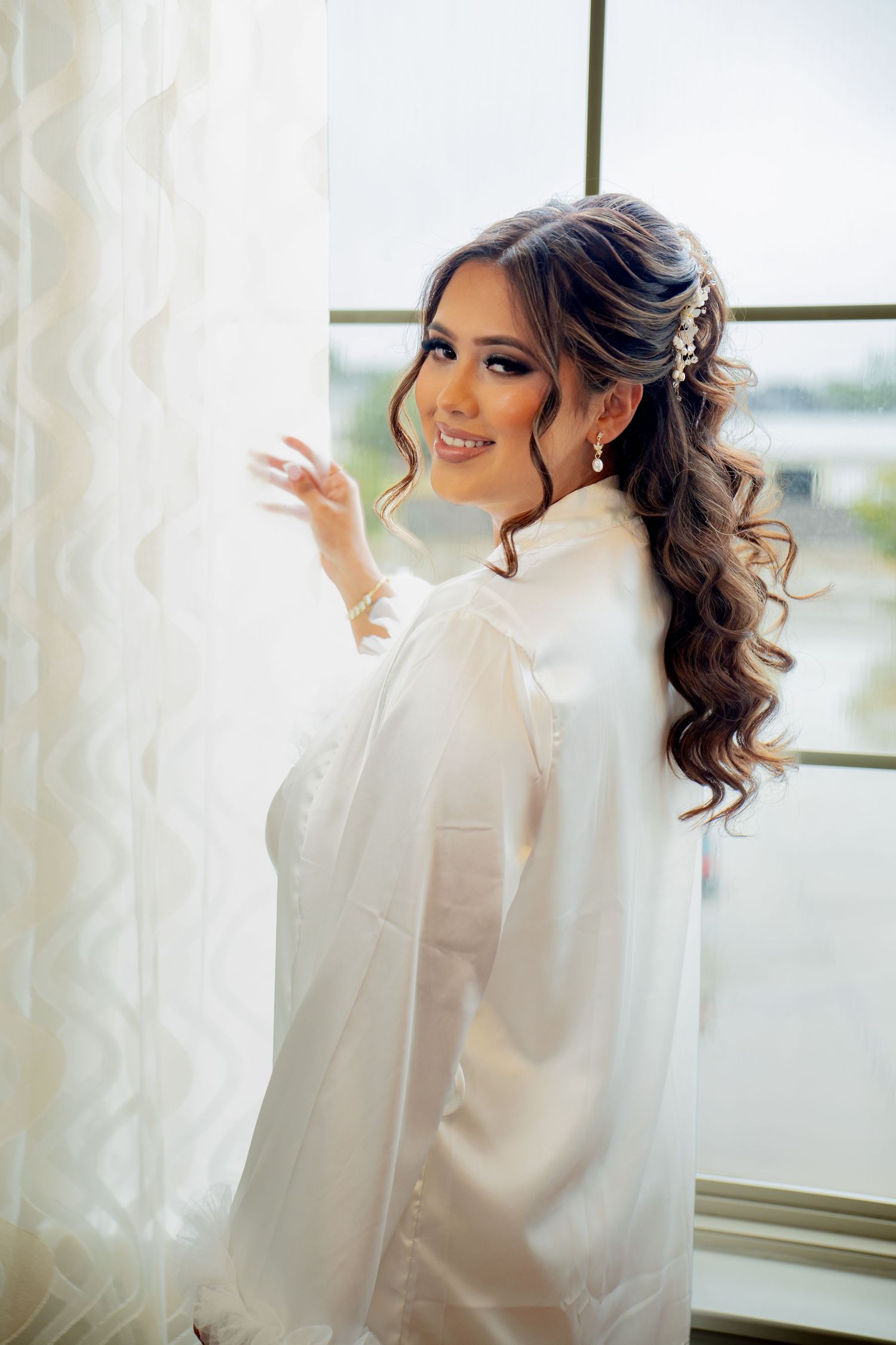 Beautiful bride in white satin robe with long curly hair and floral accessories smiles by a bright window.