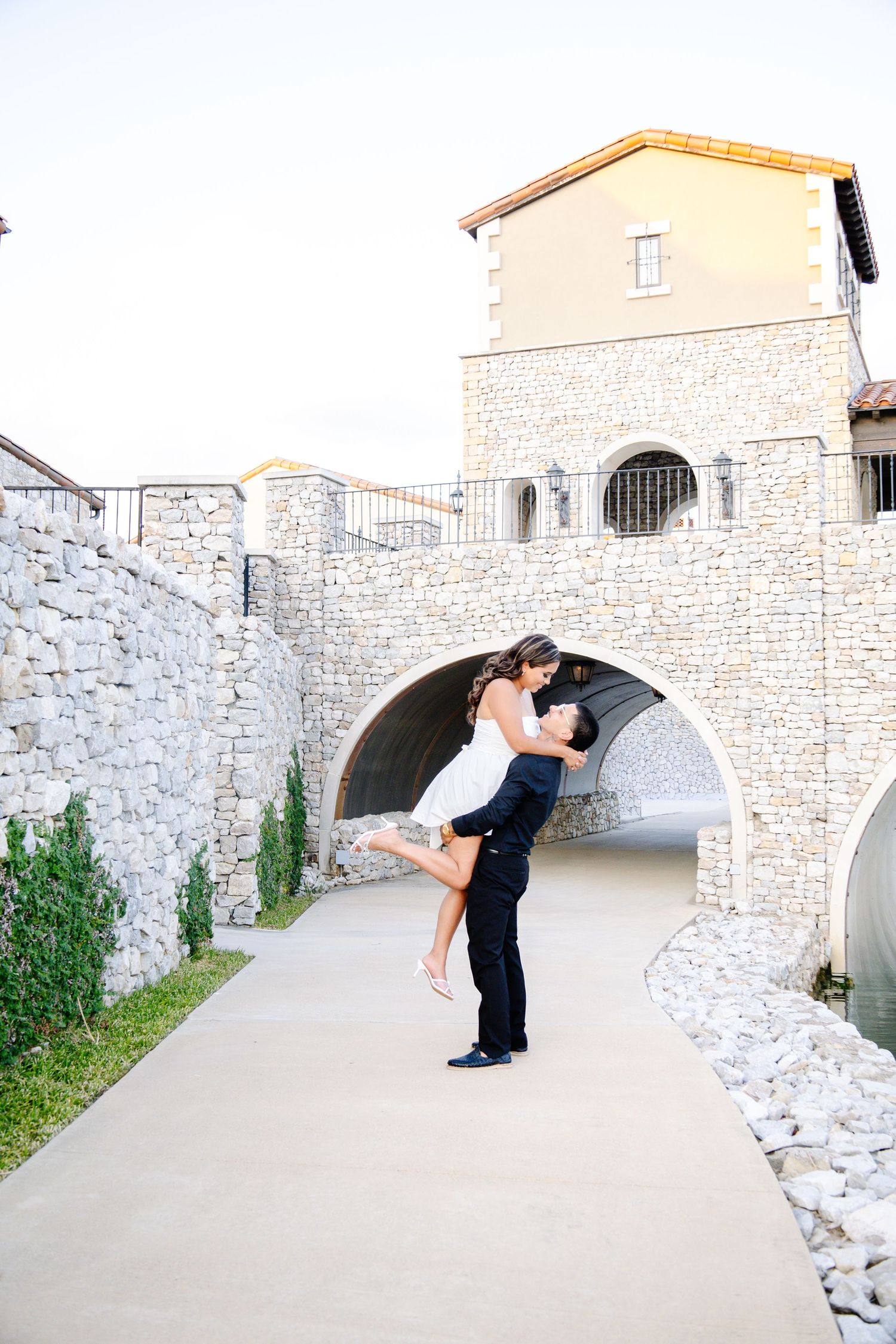 A man lifts a woman in a white dress near a stone arch walkway at a European-style venue.