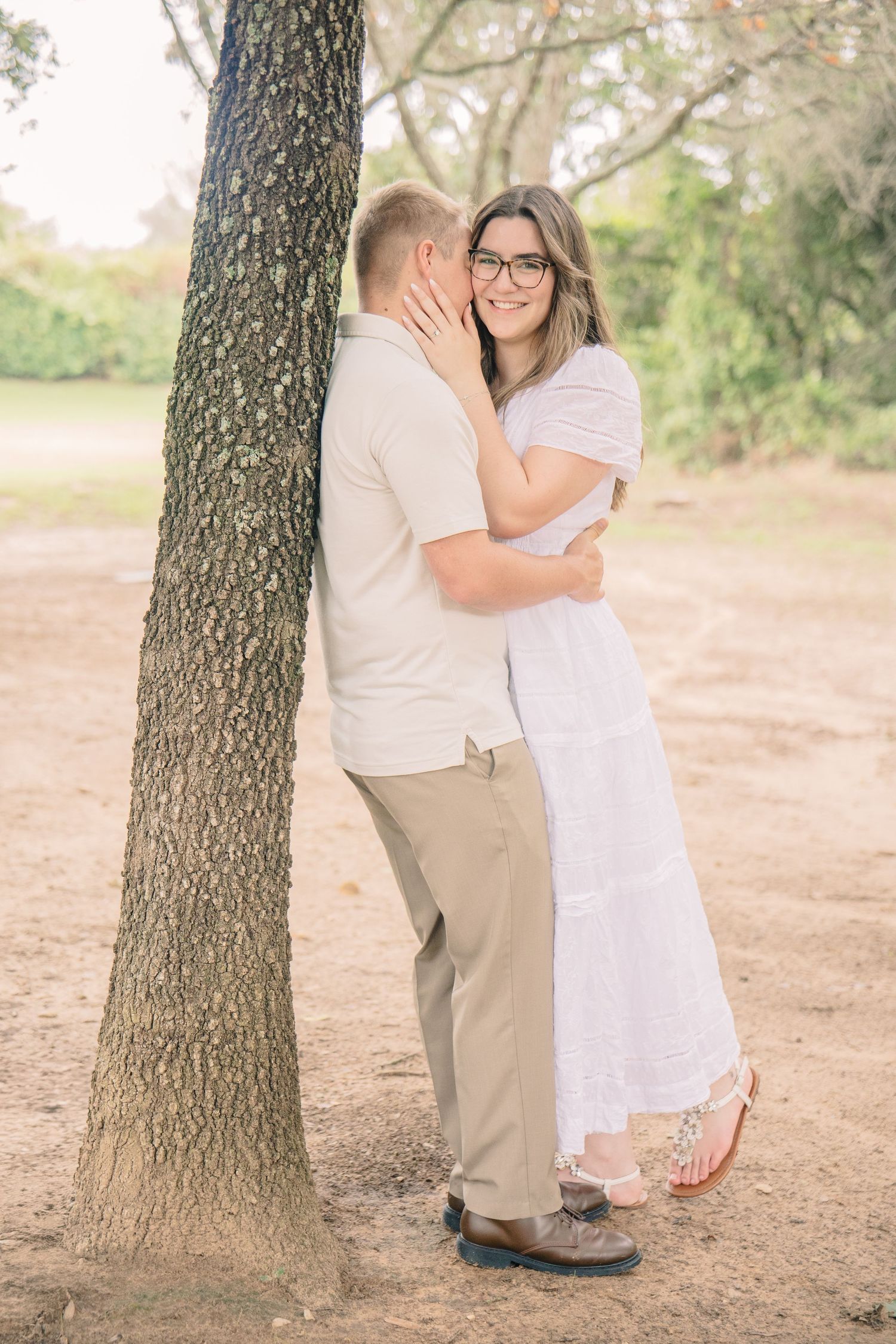 Couple embracing by a tree outdoors, woman in white dress and glasses smiling, man in beige outfit, in a natural park setting.