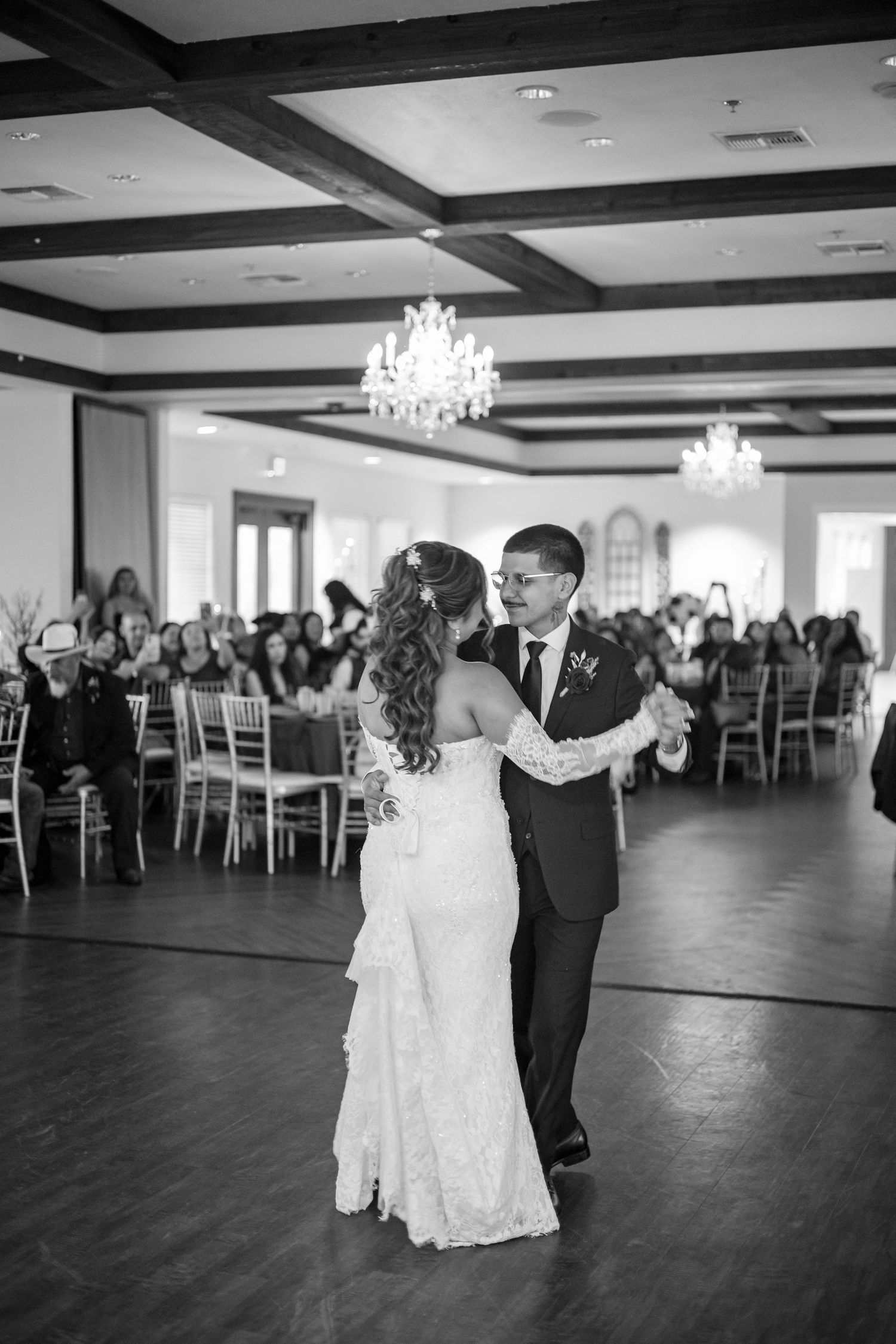 Bride and groom share first dance in elegant ballroom with chandeliers and wedding guests.