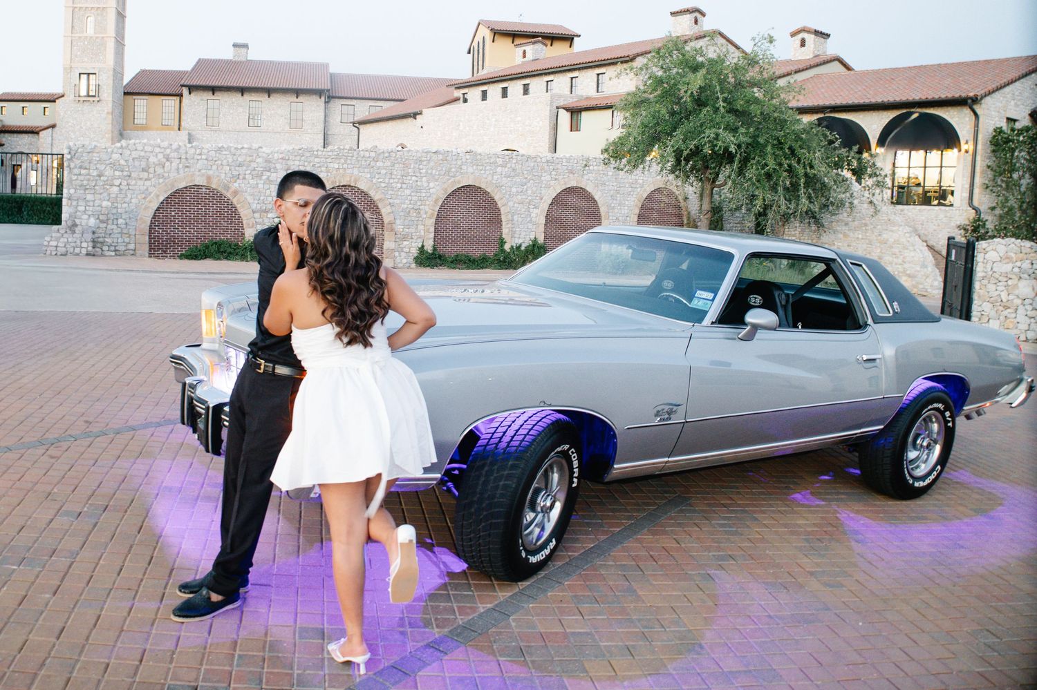 Couple sharing a romantic moment beside a classic silver muscle car with purple lighting at an elegant venue.