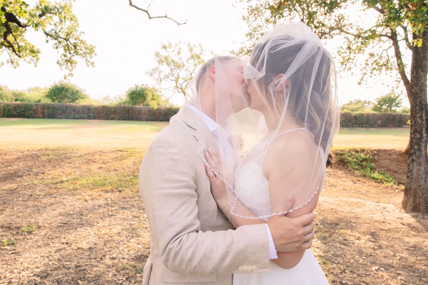 Bride and groom share a romantic kiss under a sheer veil in a sunlit outdoor field surrounded by trees on their wedding day.