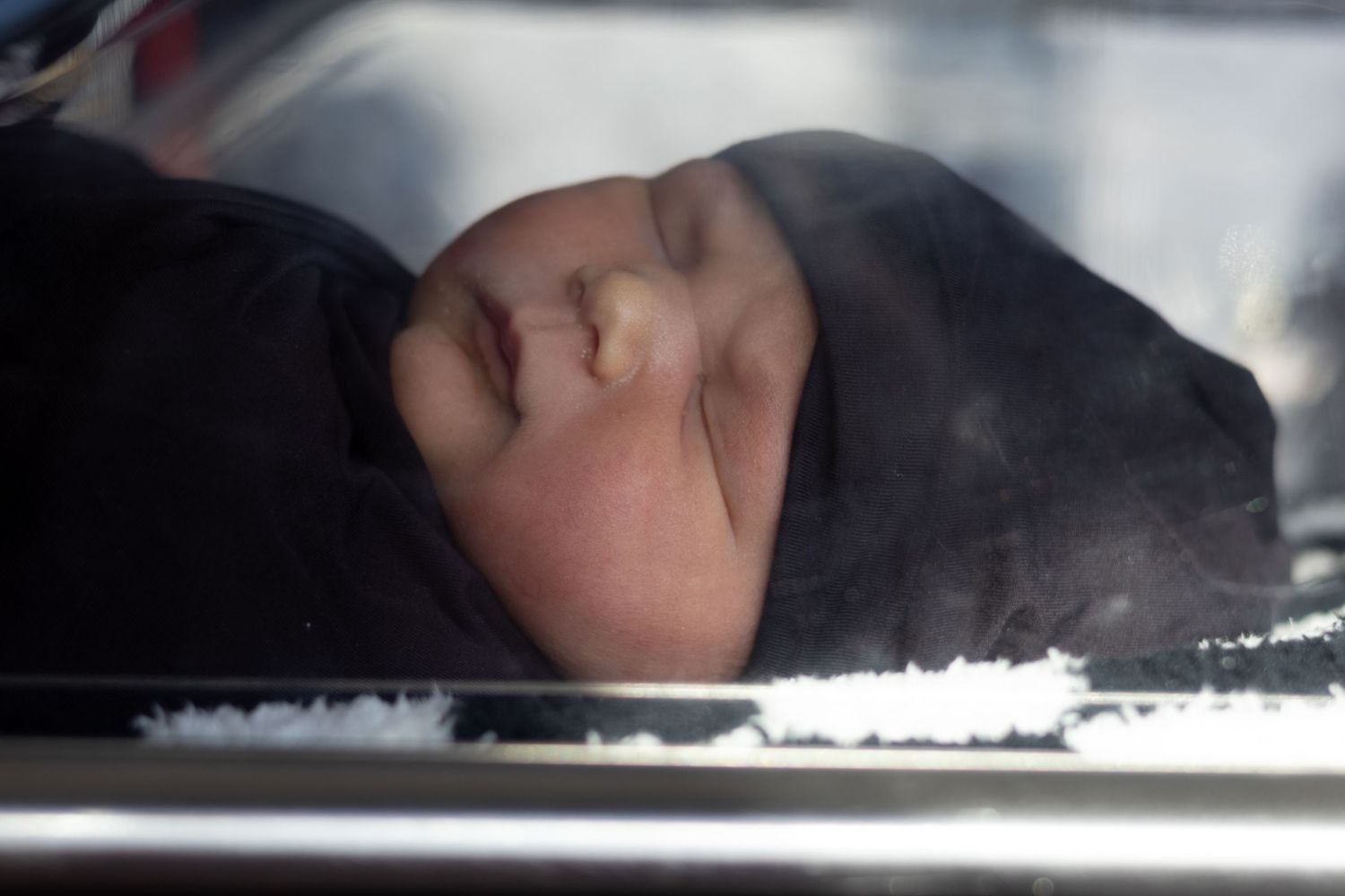 Newborn baby sleeping peacefully in a hospital bassinet wrapped in dark blanket.