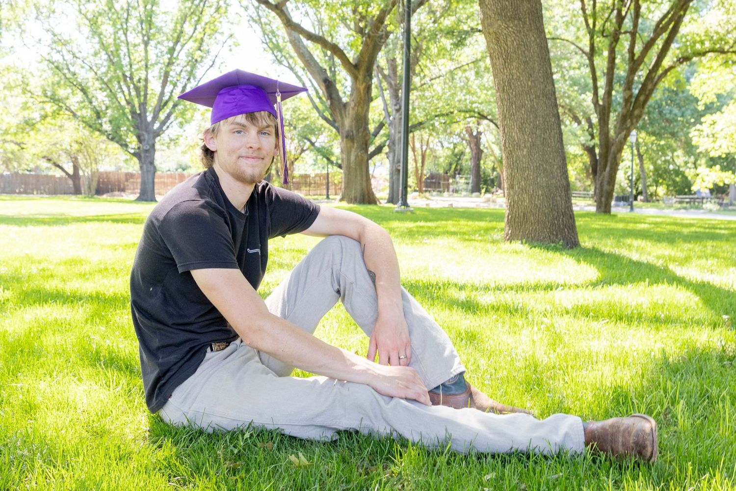 Young man wearing purple graduation cap sits casually on green grass beneath shade trees.