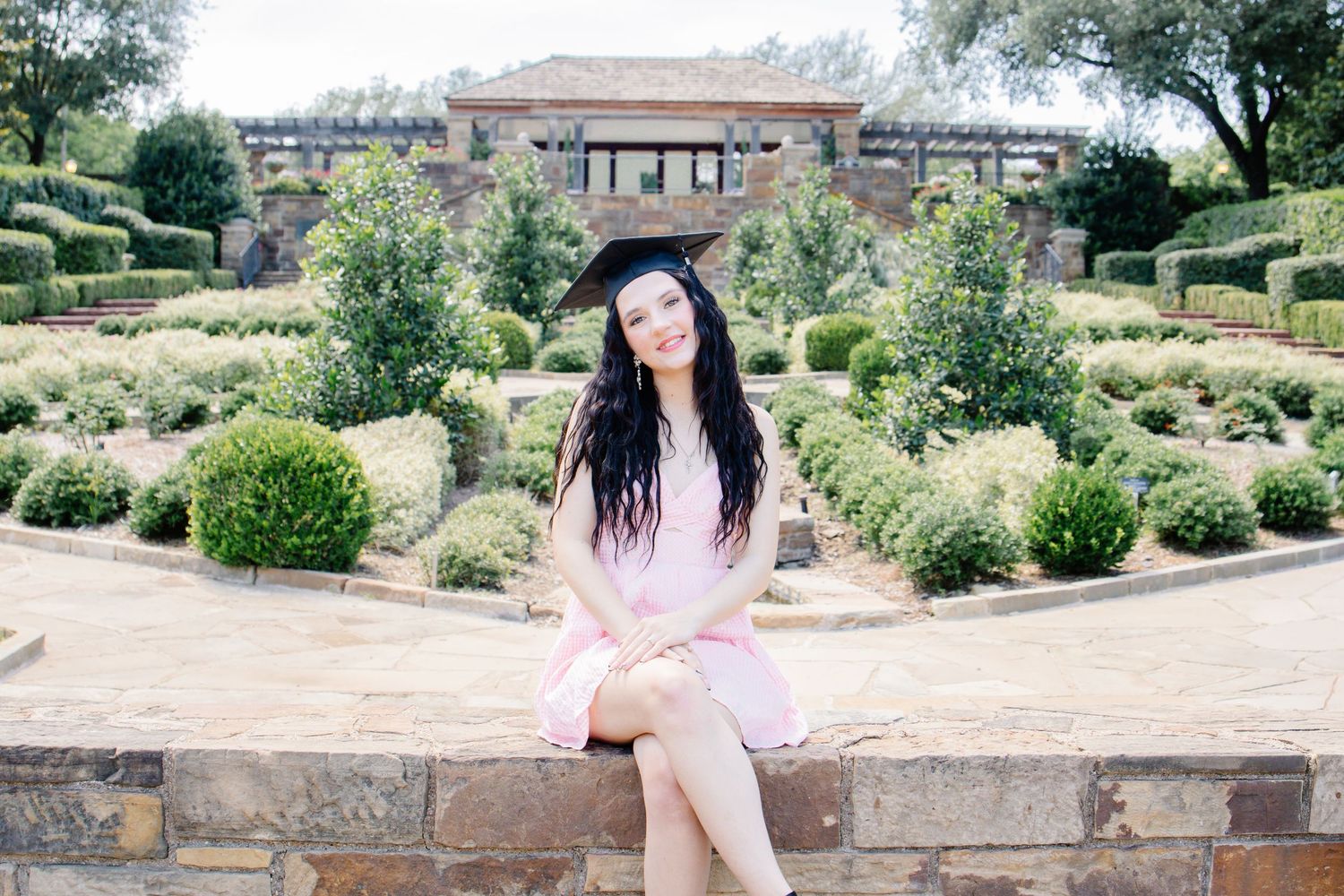 A young woman in a pink dress and graduation cap sits on a stone wall in a formal garden.