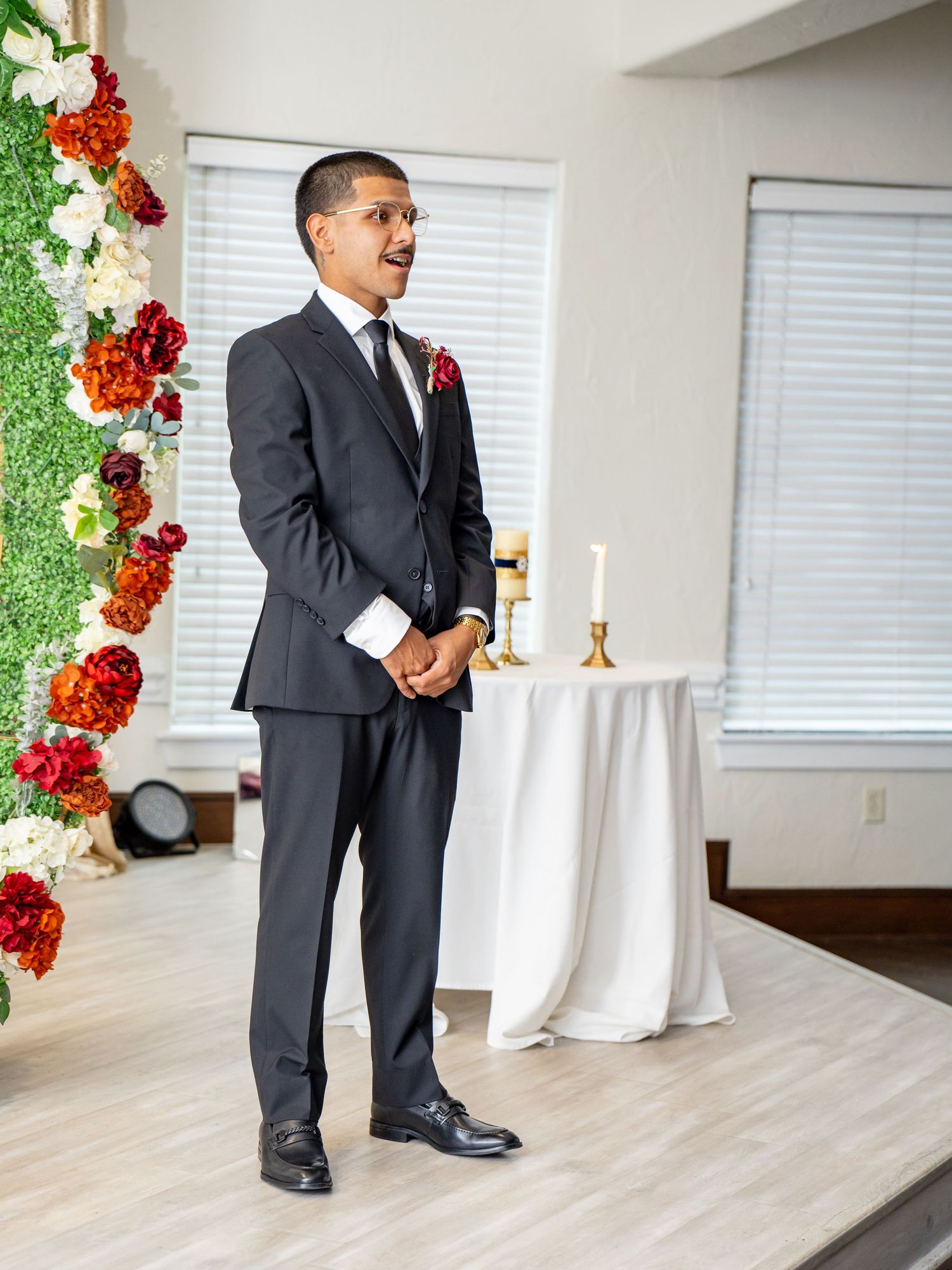 Groom in dark suit with red boutonniere stands at floral wedding altar awaiting bride.