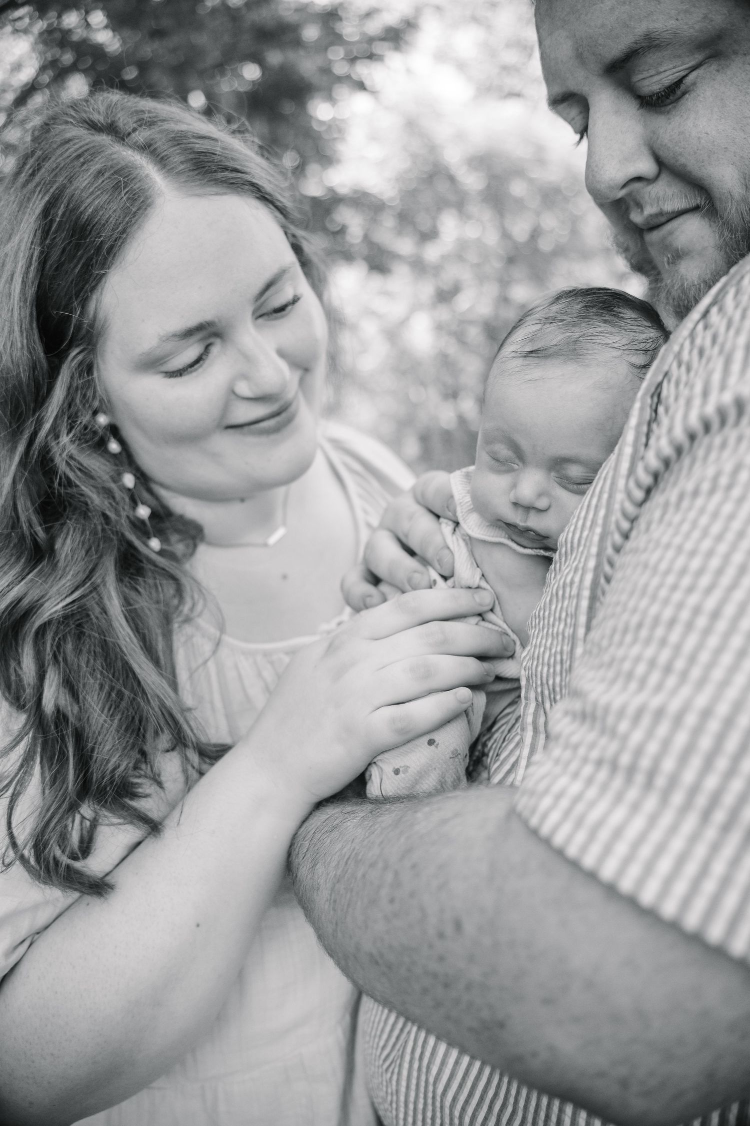 Black and white photo of parents lovingly gazing at their sleeping newborn baby outdoors.