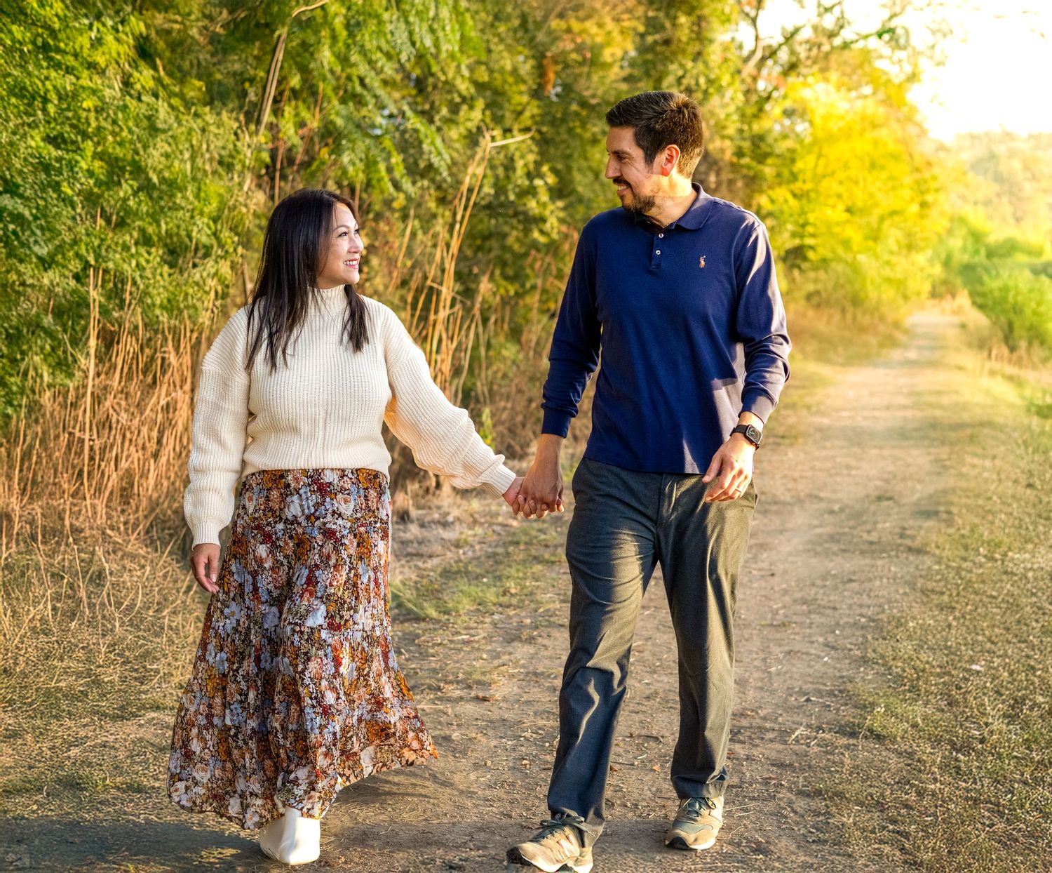 Happy couple walking hand in hand on dirt path surrounded by golden foliage.