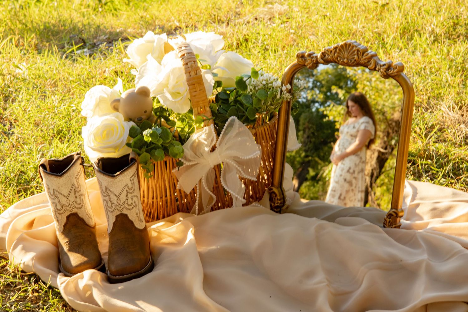 Rustic maternity flat lay with cowboy boots, white roses in a basket, and a small mirror reflecting a pregnant woman outdoors.