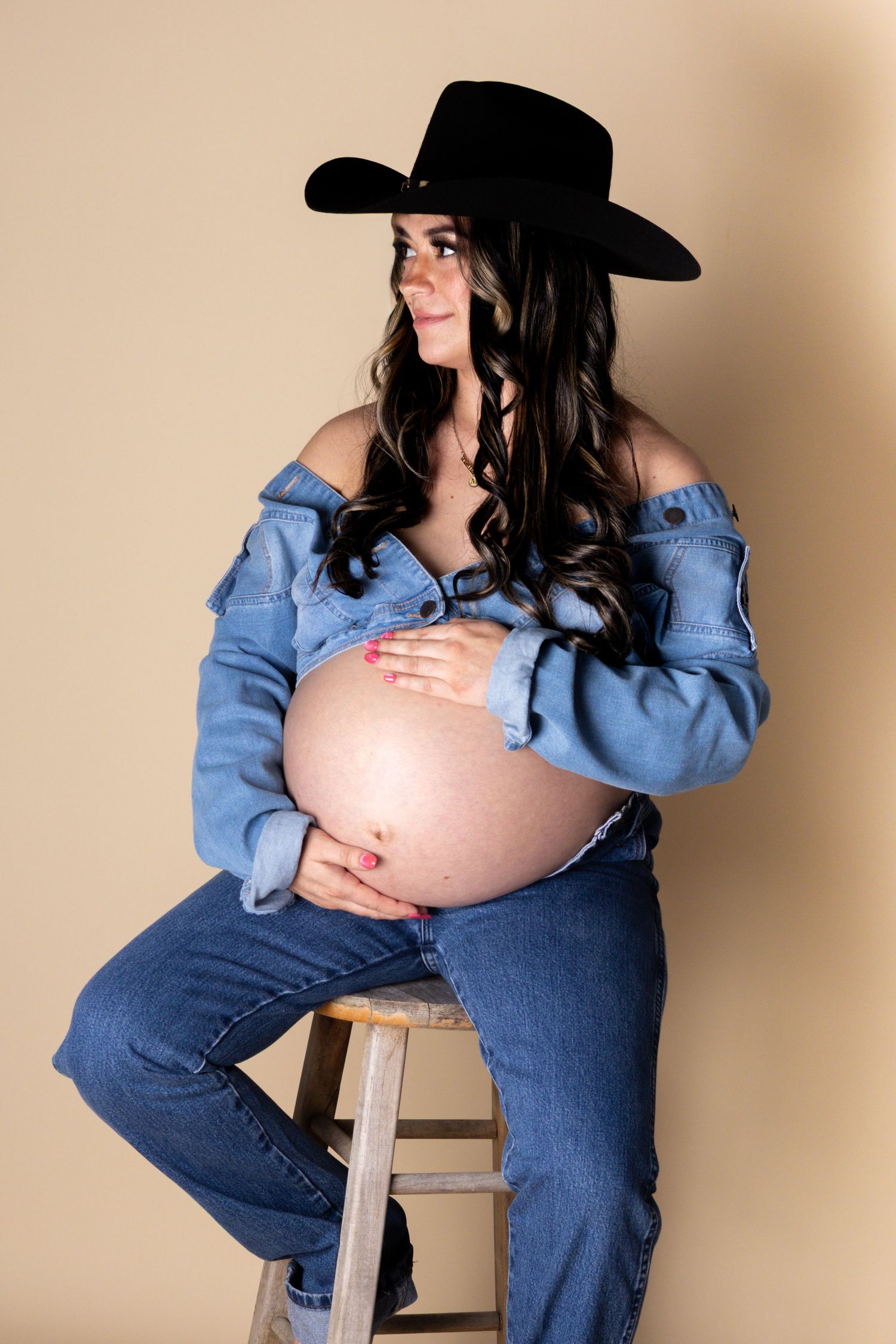 Smiling pregnant woman in cowboy hat and denim outfit sitting on stool, cradling belly.