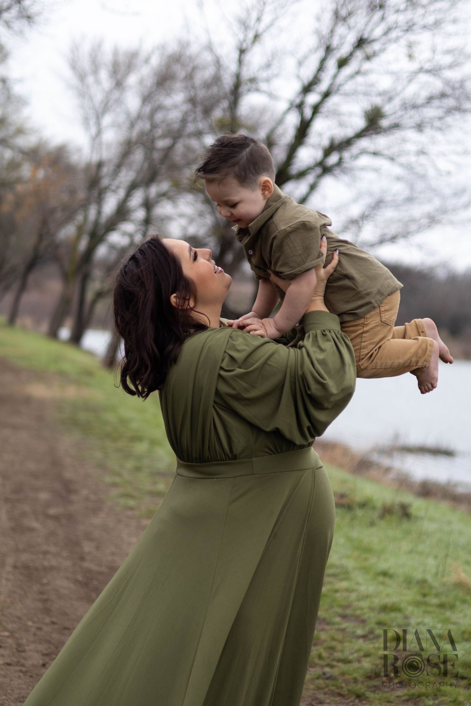 Mother in olive green maternity dress lifts smiling baby boy overhead near a lake during photo session.