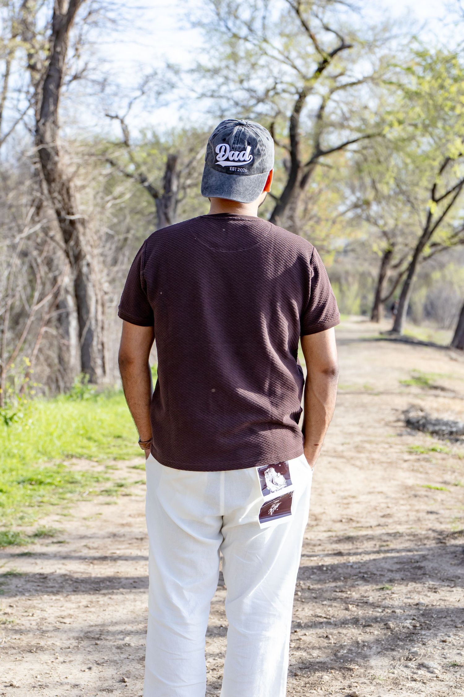 Man wearing a Dad cap walking away on a sunny tree-lined path with an ultrasound photo in his pocket.