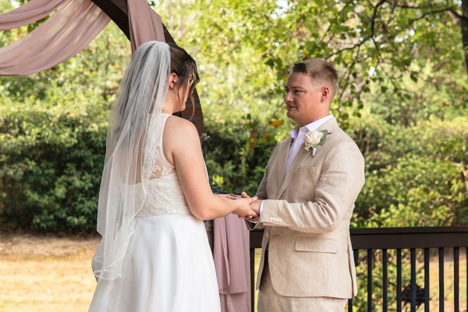 A bride in a white dress and veil holds hands with a groom in a tan suit during an outdoor wedding ceremony.