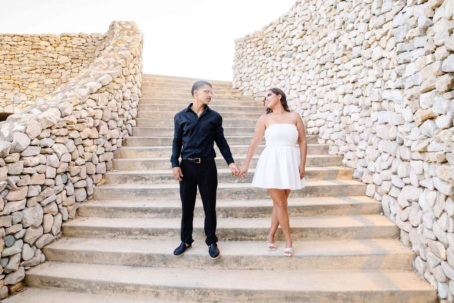 Couple holding hands on stone staircase, man in black outfit and woman in white dress, outdoor engagement photo.
