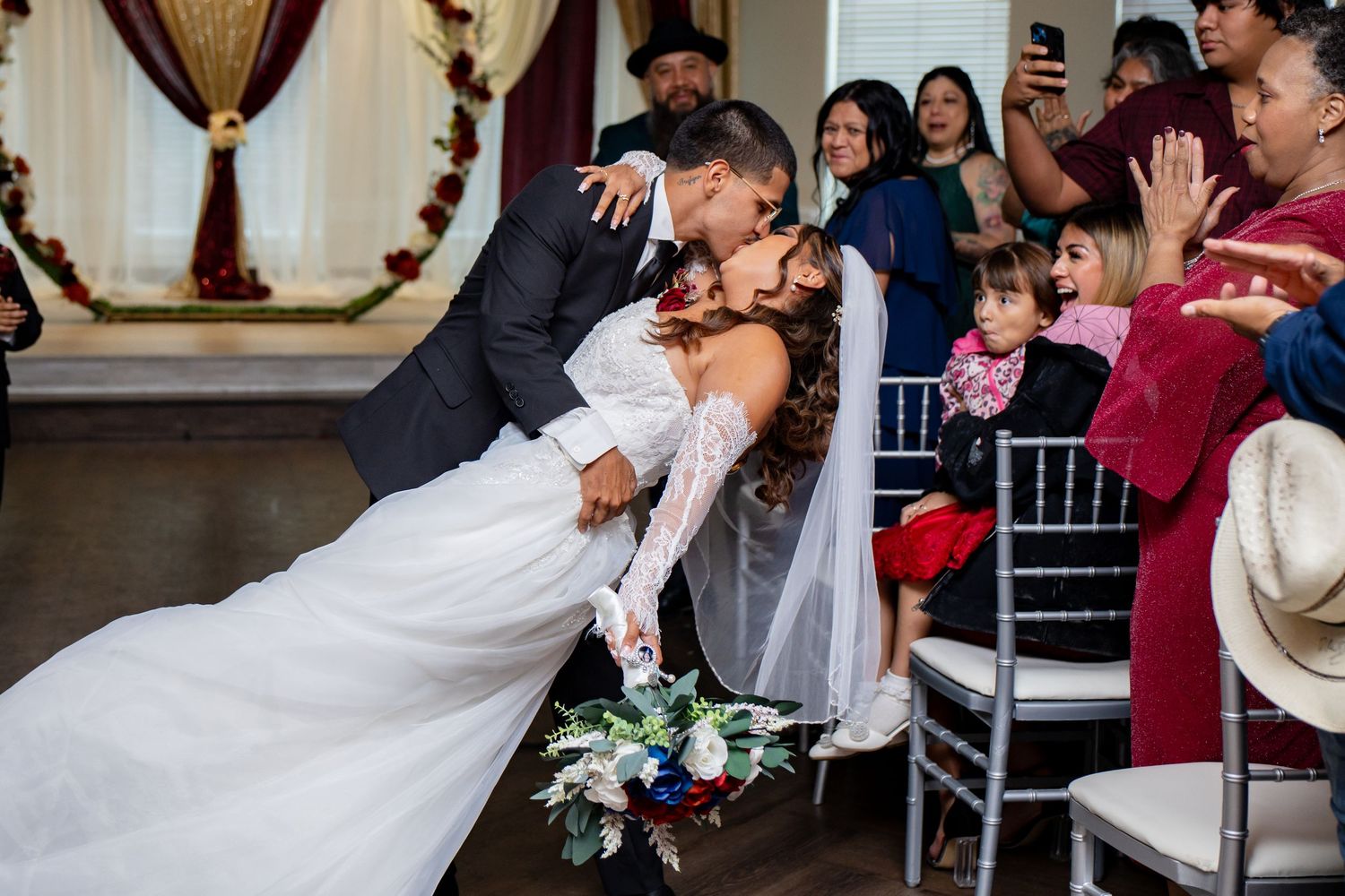 Groom dips bride for kiss at wedding ceremony aisle surrounded by guests.