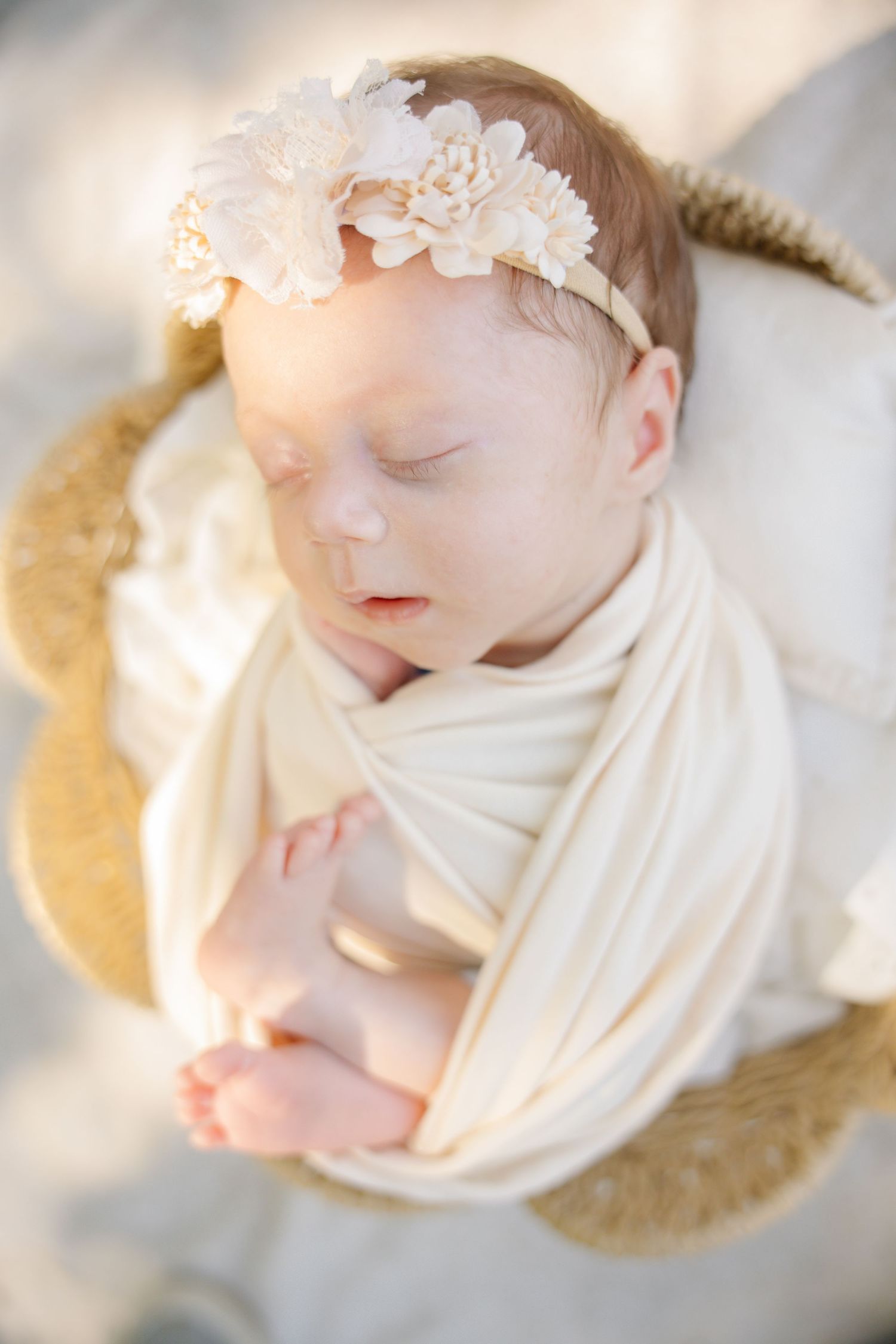 Sleeping newborn baby girl swaddled in cream wrap with floral headband in a wicker basket.