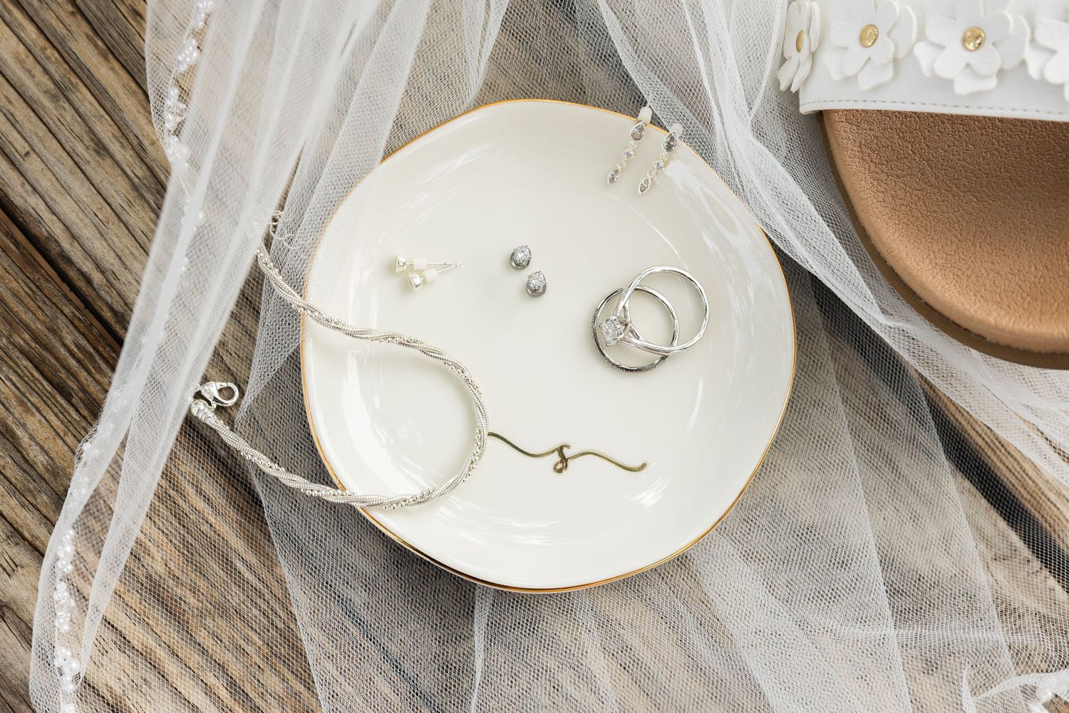 White ceramic dish holding wedding rings, pearl earrings, and a delicate necklace on a wooden surface with bridal veil.