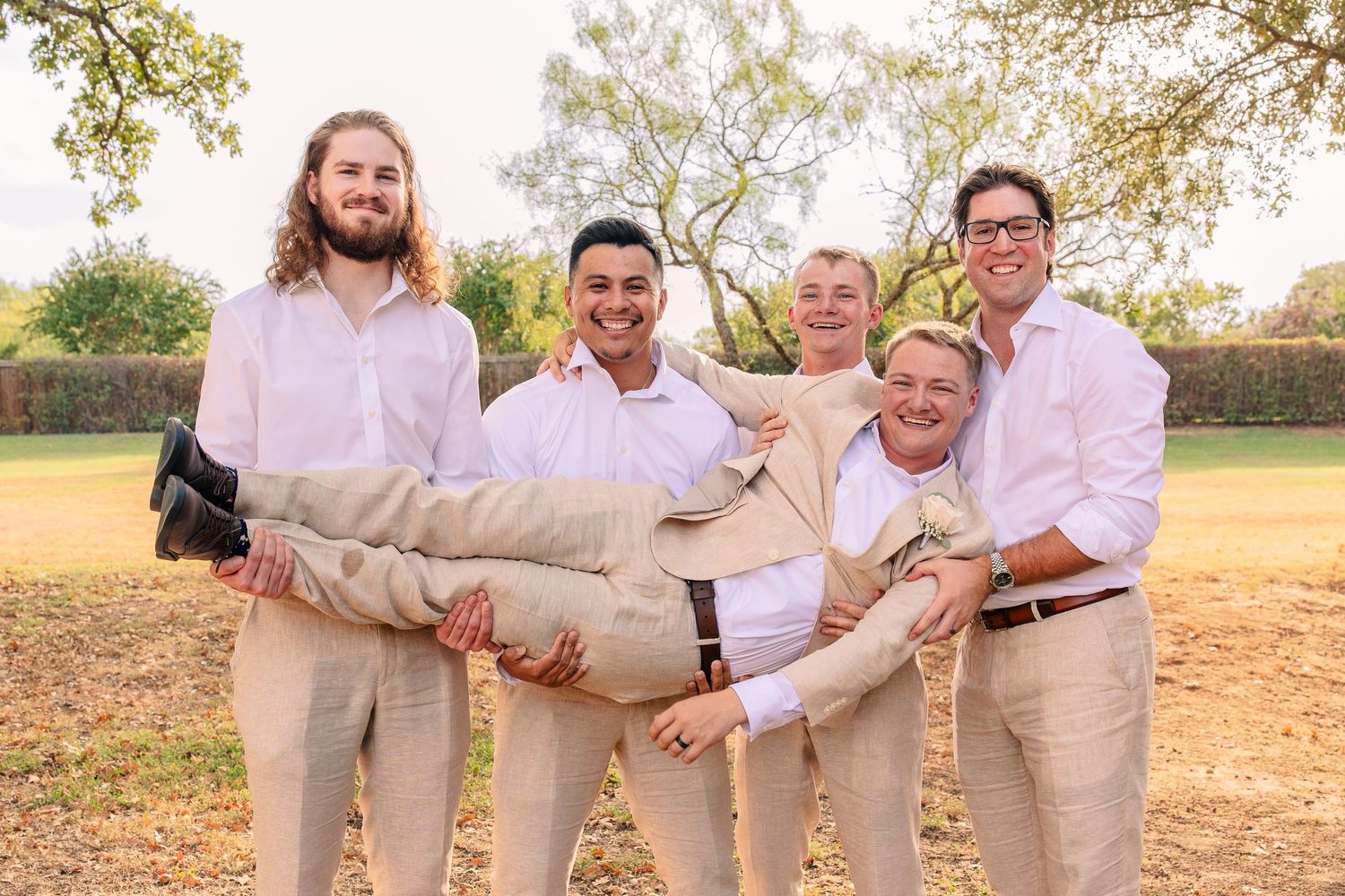Four groomsmen in white shirts and khaki pants playfully lift the laughing groom in a candid outdoor wedding photo.