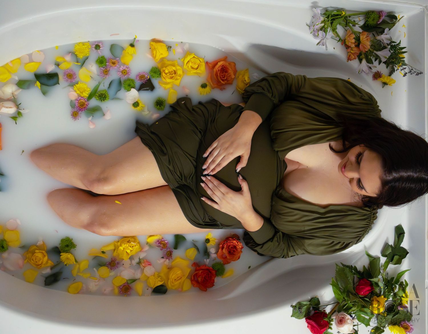Overhead view of pregnant woman in olive wrap dress lying in floral milk bath with colorful roses.
