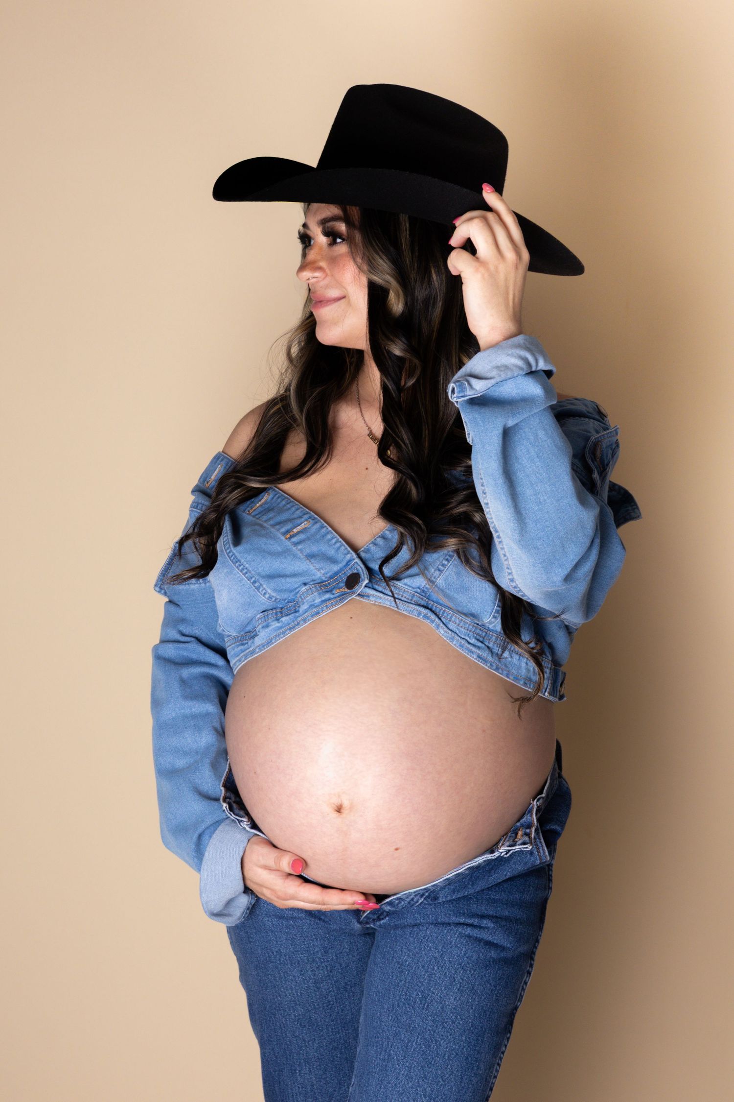 Pregnant woman in black cowboy hat and denim jacket touching hat brim, showing bare belly.