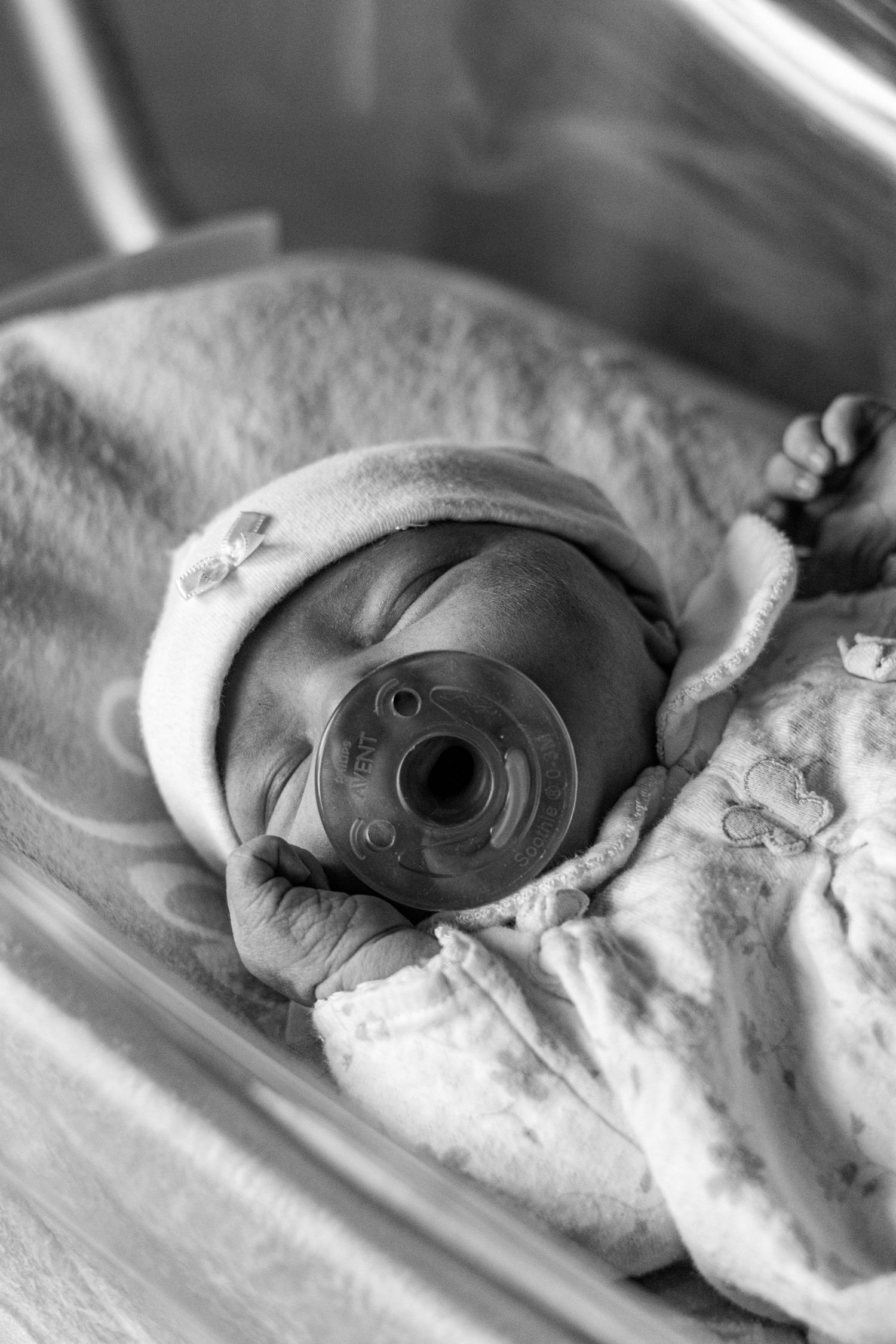 Black and white photo of a swaddled newborn baby sleeping with a pacifier in hospital bassinet.